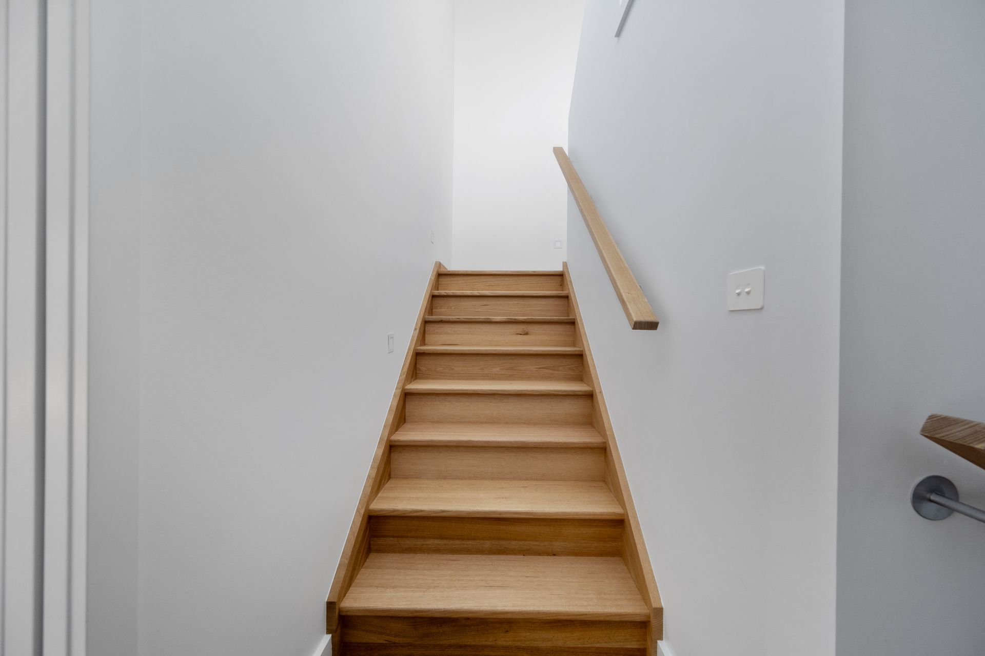 Wooden stairs leading upward between white walls, with a wooden handrail on the right.