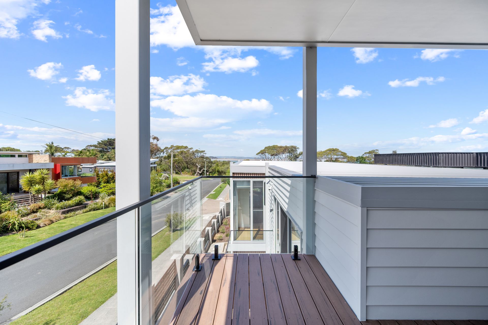 Balcony view of a modern home overlooking a residential area and blue sky with clouds. White and grey accents with a wooden deck.