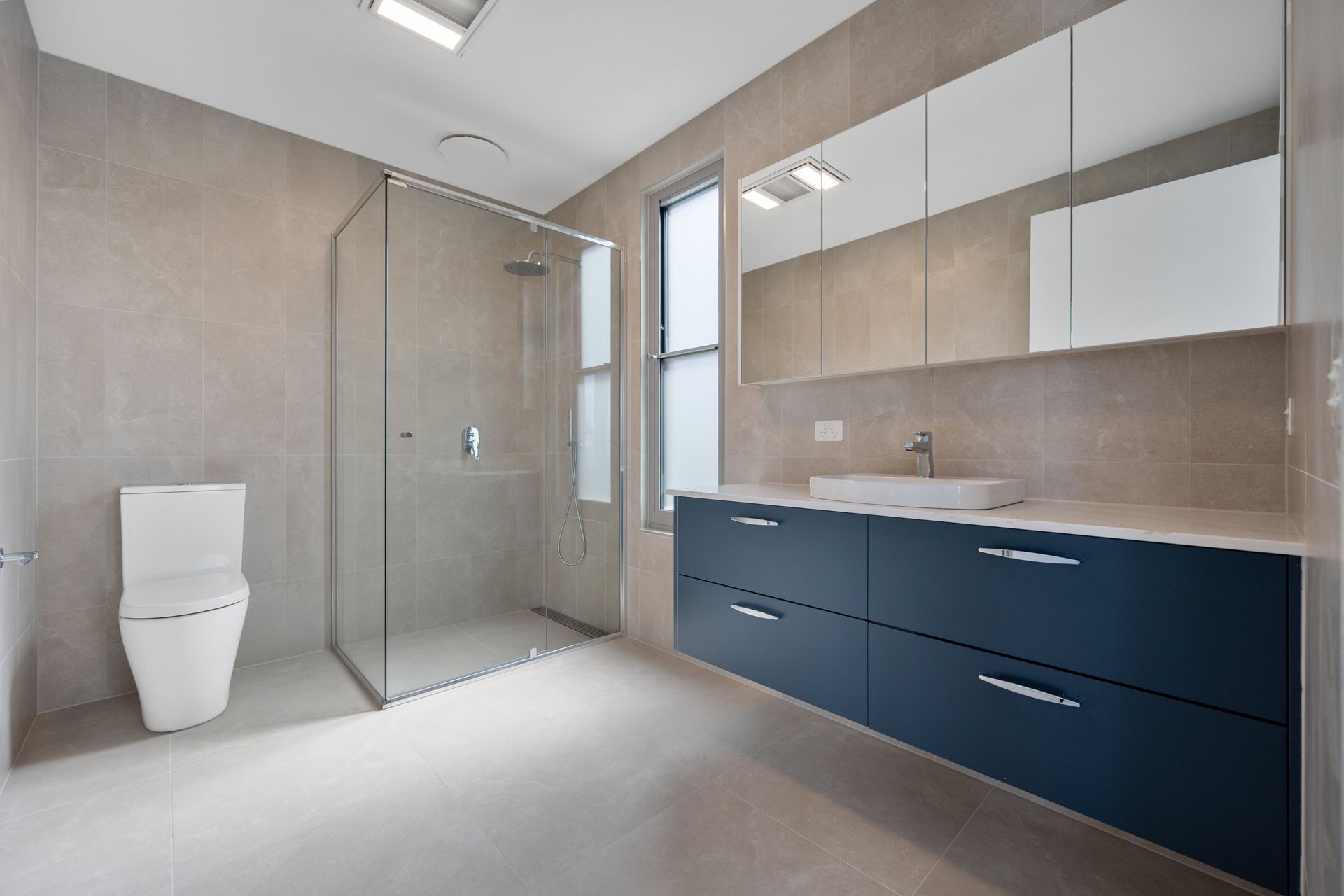Modern bathroom with a blue vanity, white sink, glass shower, and toilet. Beige tile covers the walls and floor.