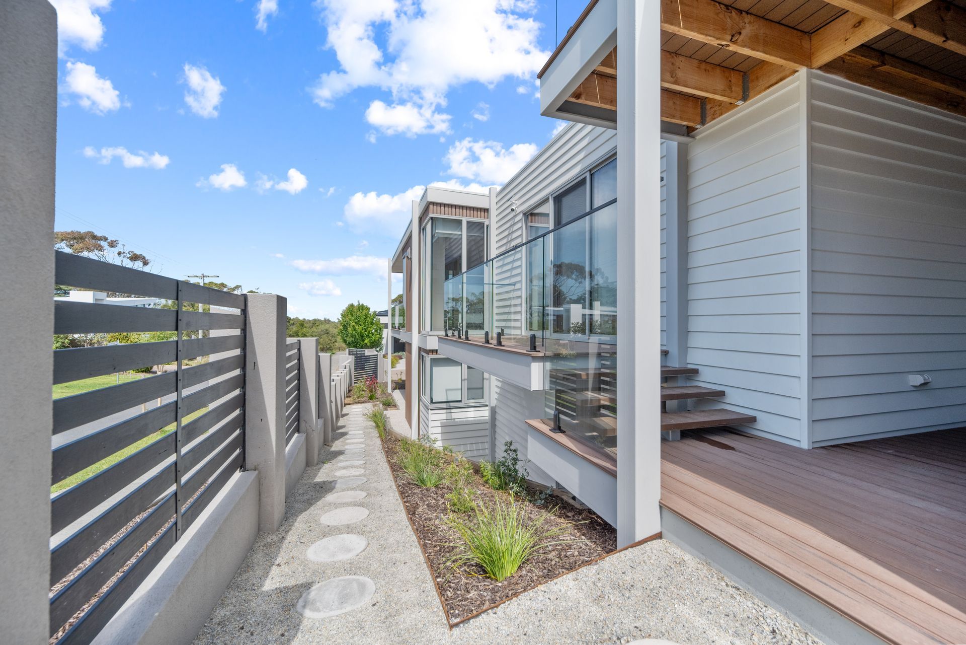 Modern home exterior with gray and white siding, wooden deck, and glass balcony railing. Pathway lined with a gray fence.