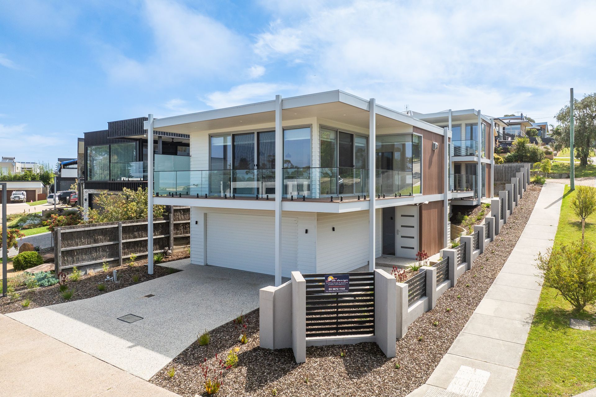 Modern two-story home with a white exterior, glass balconies, and a driveway. It is located on a sloped street with other houses nearby.