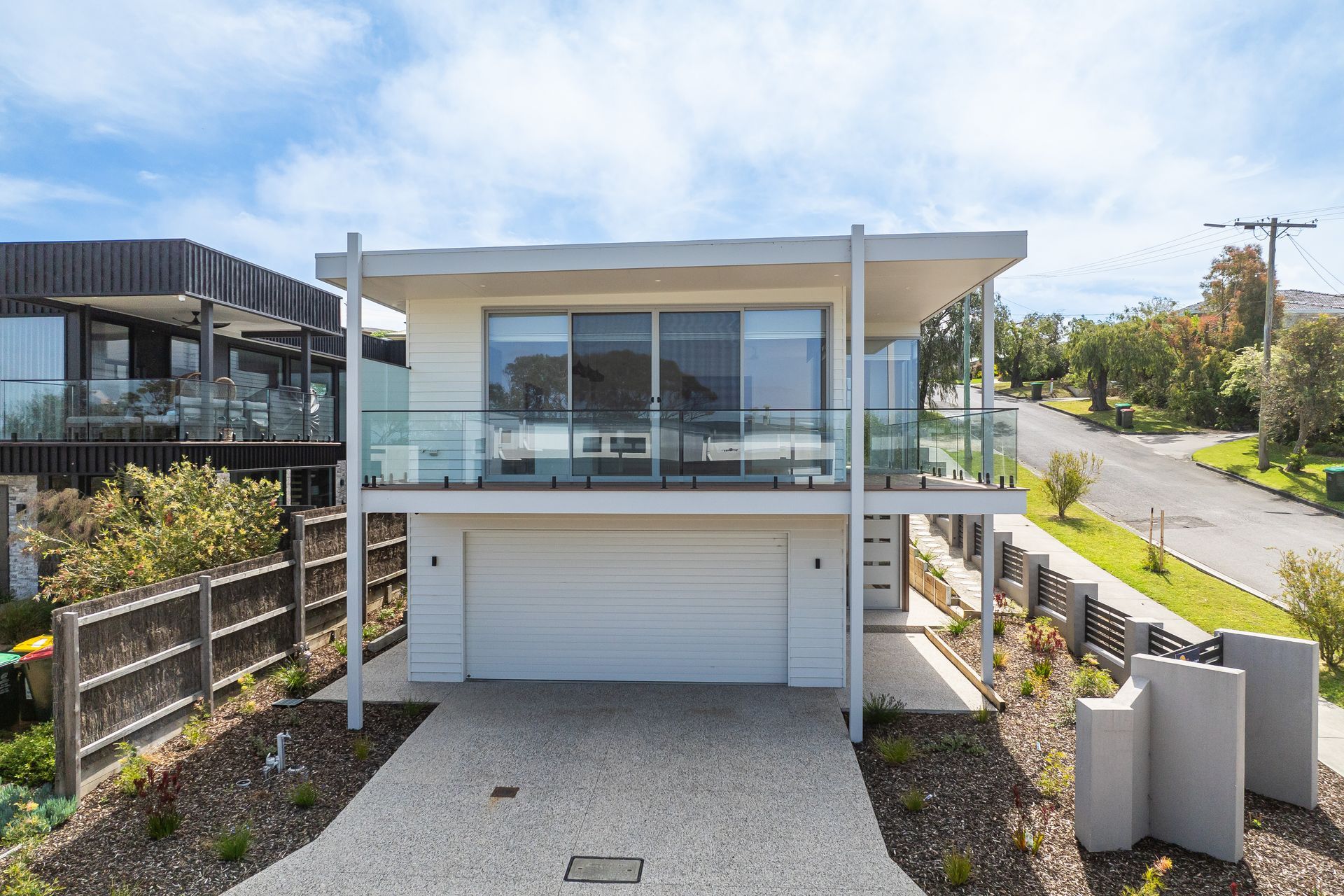 Modern, two-story house with a white facade, glass balconies, and a garage. Located on a hill, overlooking a street with trees.