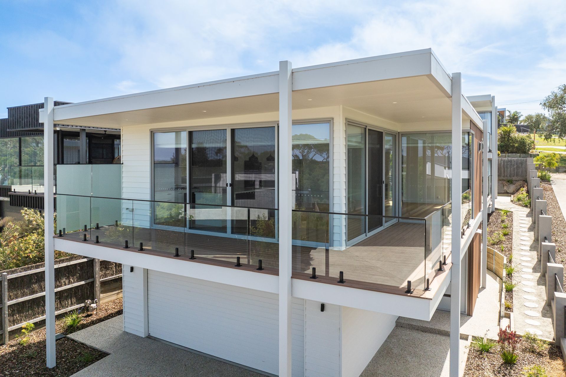 Modern white house with a deck, glass railings, and large windows, under a blue sky.