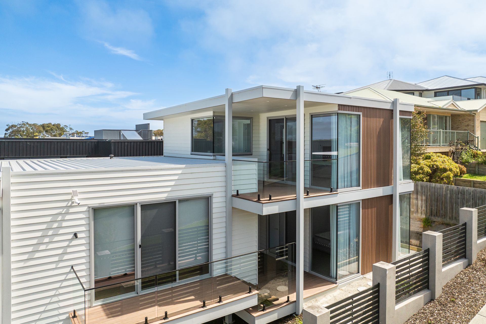 Modern two-story house with white siding and a brown accent wall.  It has balconies and is near a fence under a blue sky.