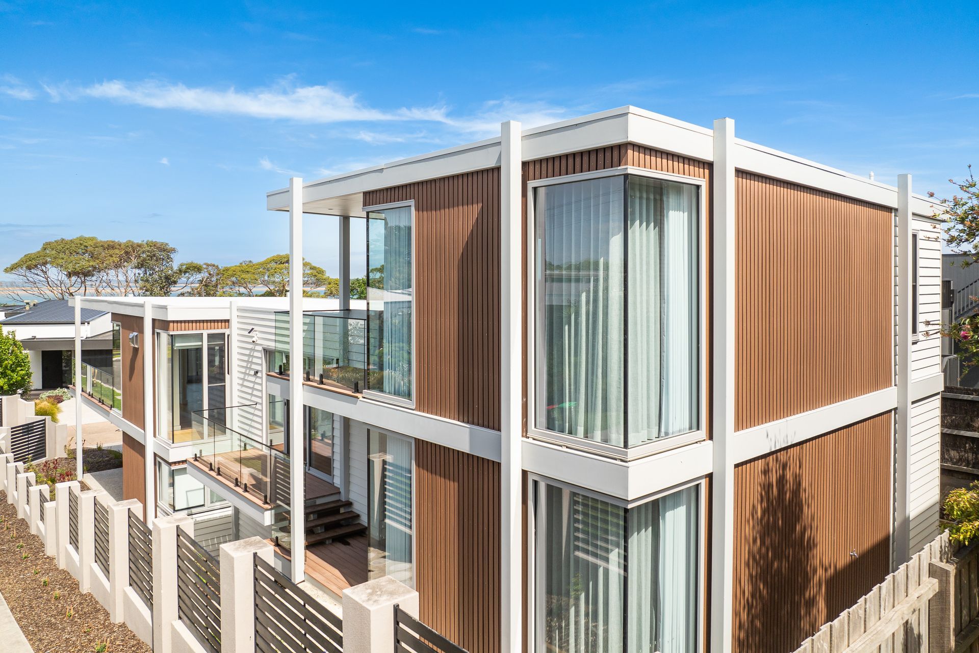 Modern two-story house with brown siding, white trim, and large windows under a blue sky. Features a balcony and fenced yard.