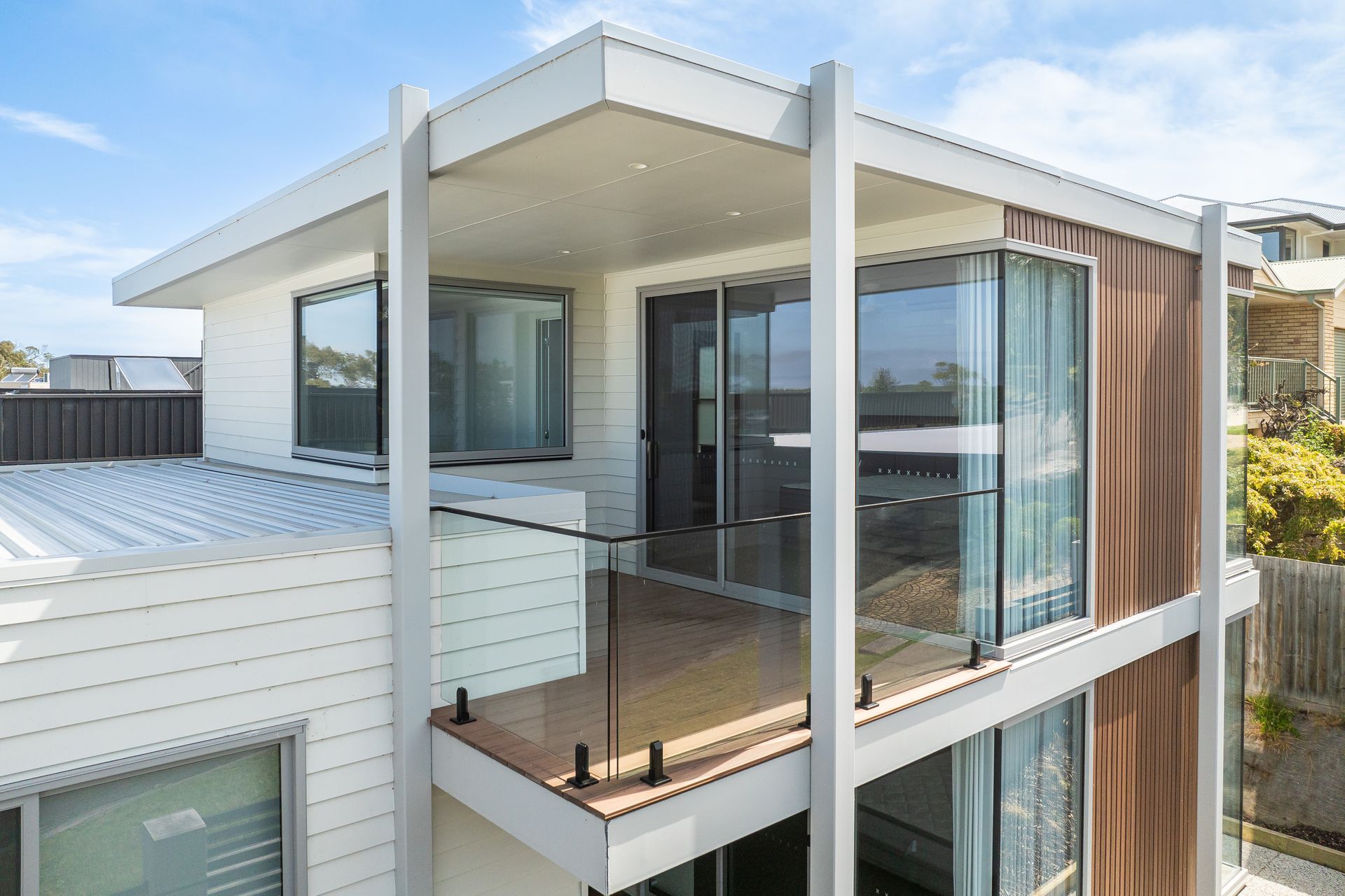 Modern two-story house with a balcony and glass railing. White siding and brown accents under a blue sky.