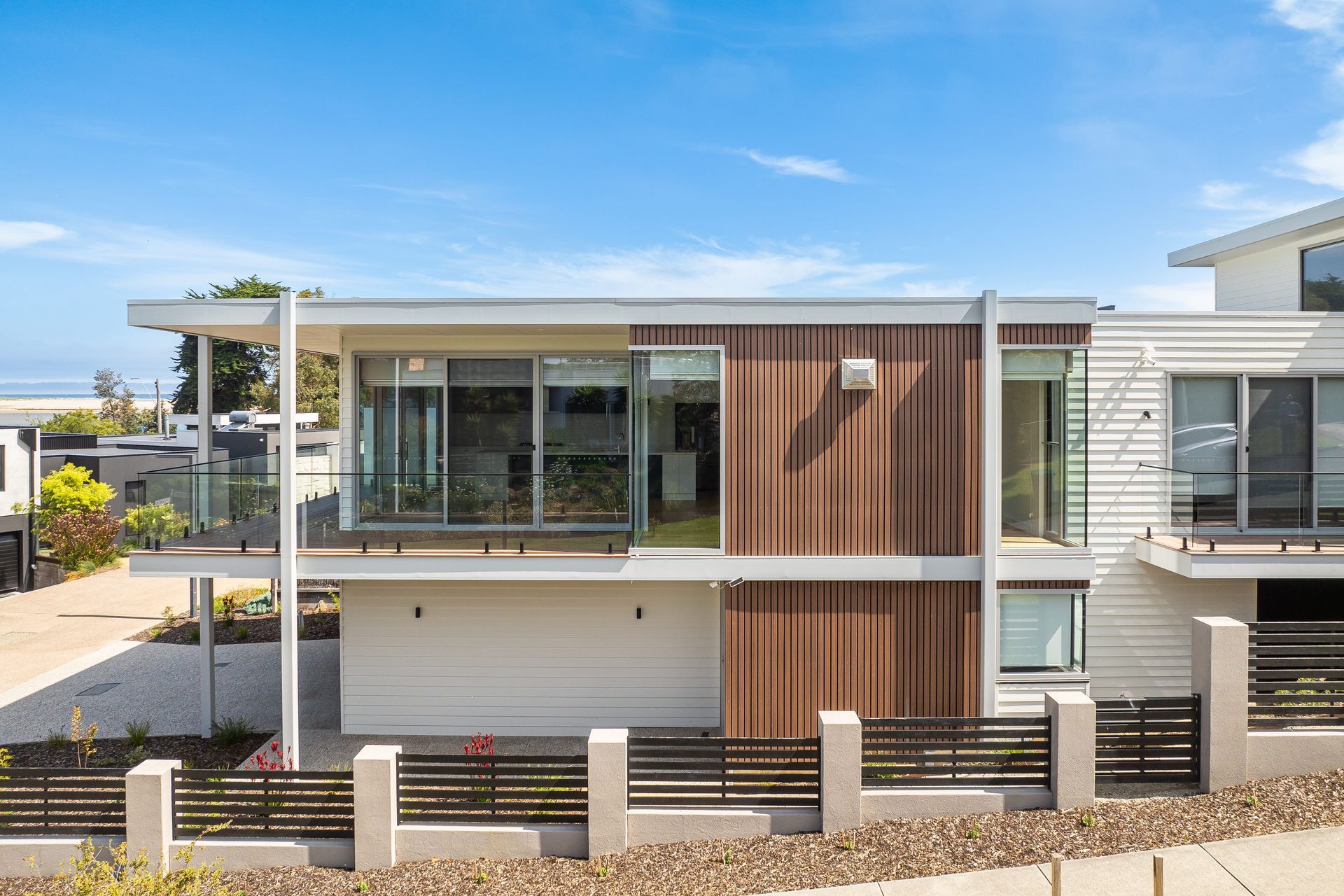 Modern two-story house with a brown wood-paneled section.  Large windows and a balcony with ocean view. Gray fence in front.