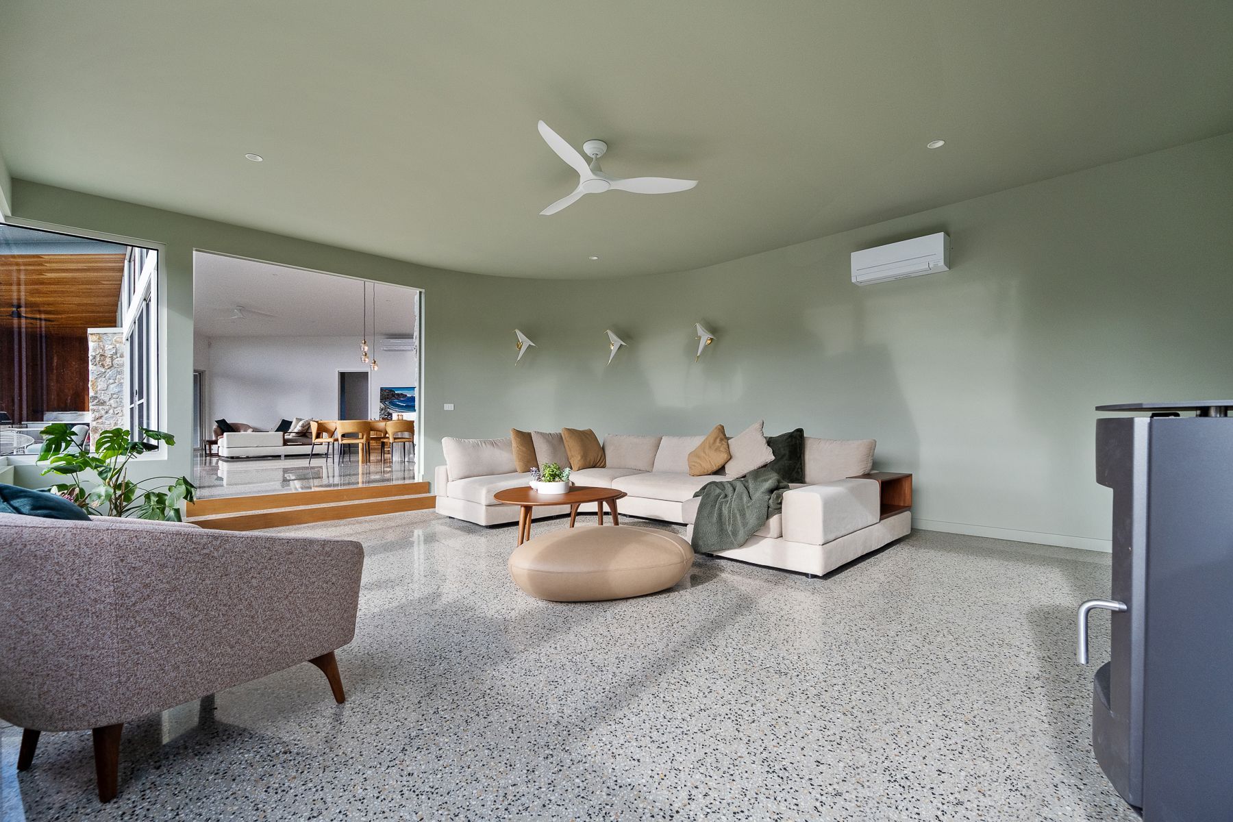 Spacious living room with light green walls, terrazzo floors, and a cream-colored sectional sofa. A round ottoman and patterned armchair sit in the foreground.