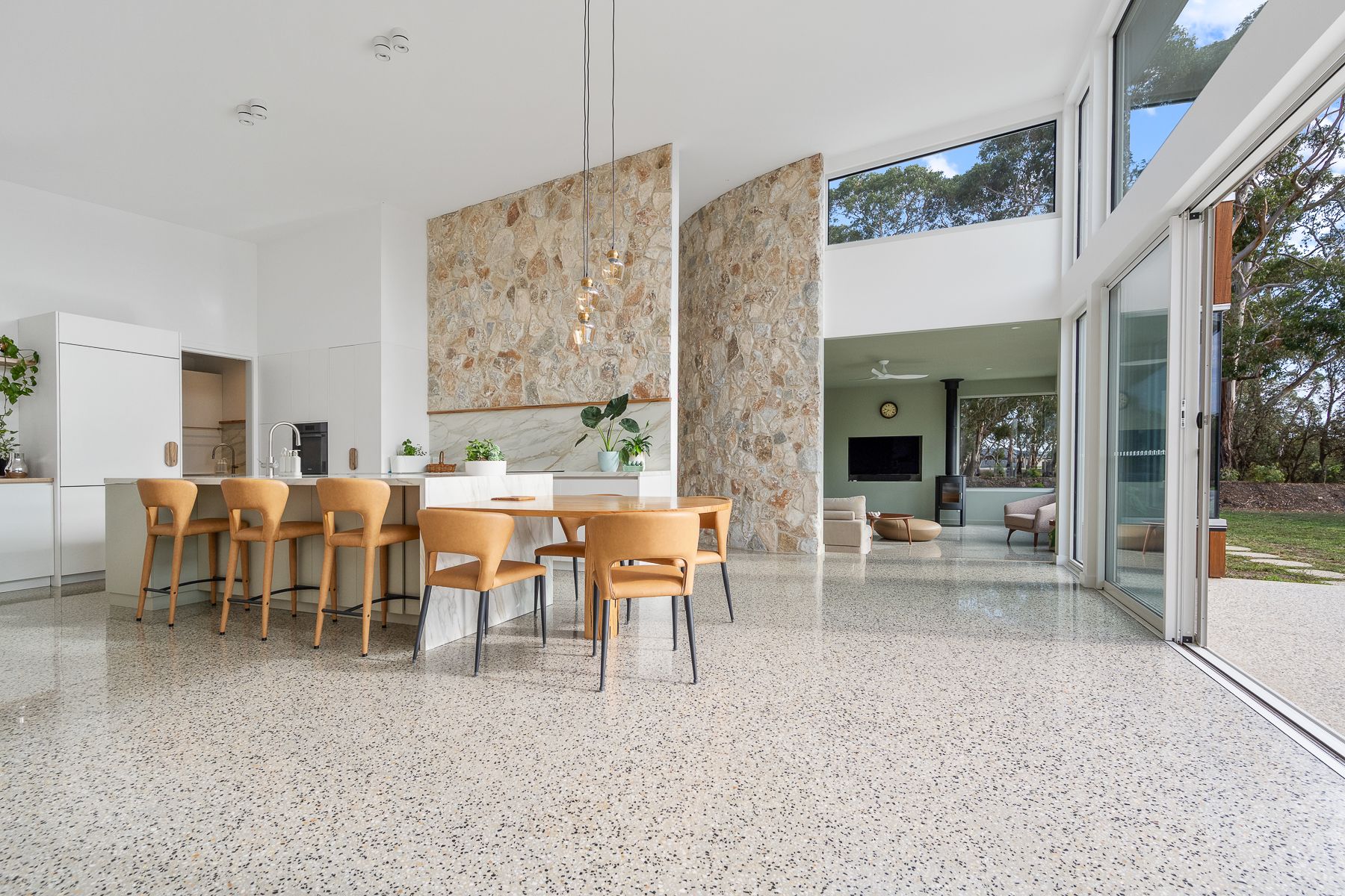 Spacious, modern kitchen and dining area with large windows, stone accents, and light wood furniture. The floor is speckled.