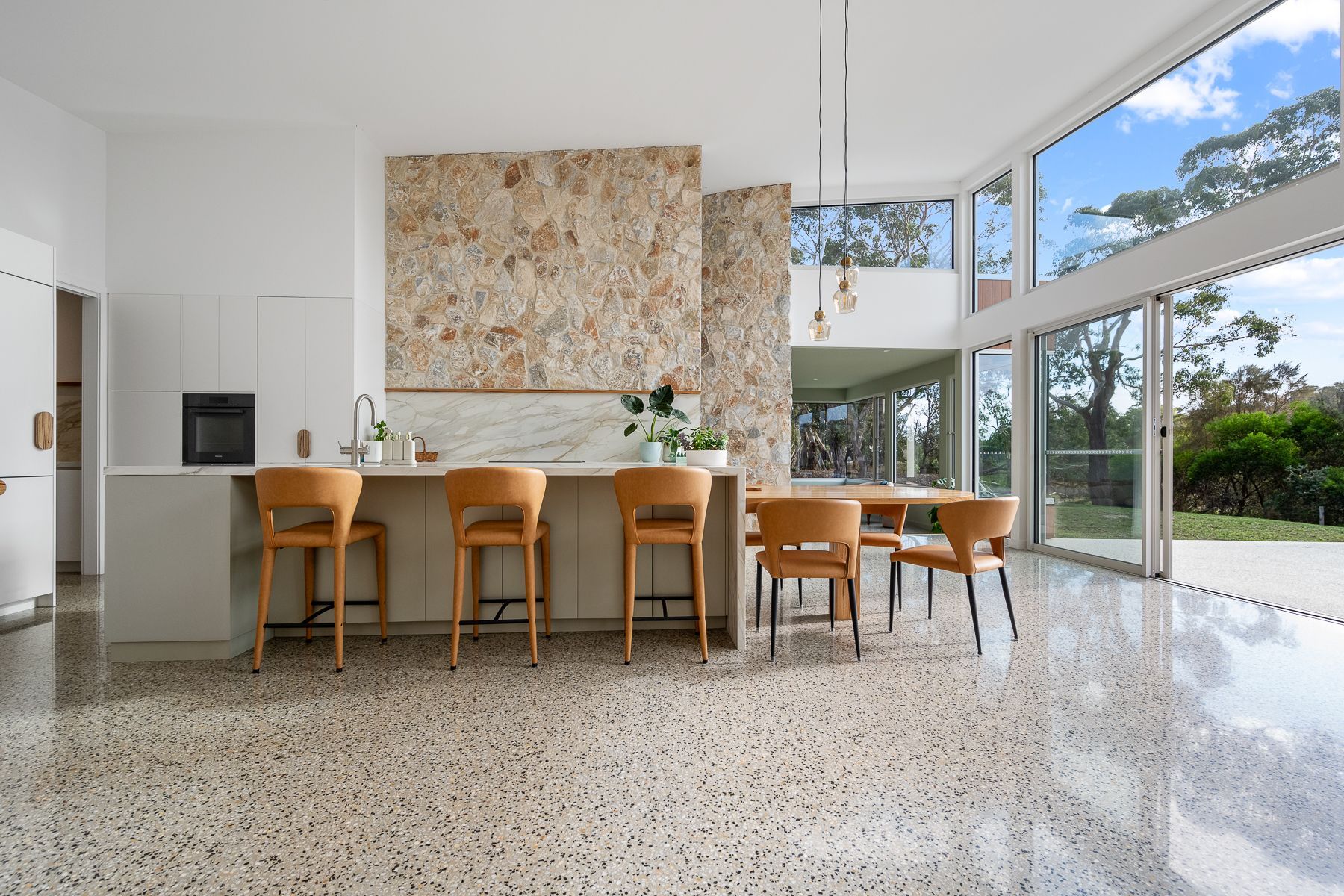 Modern kitchen with a stone accent wall, light wood barstools and dining chairs, terrazzo floor, and large windows overlooking a lush outdoor view.
