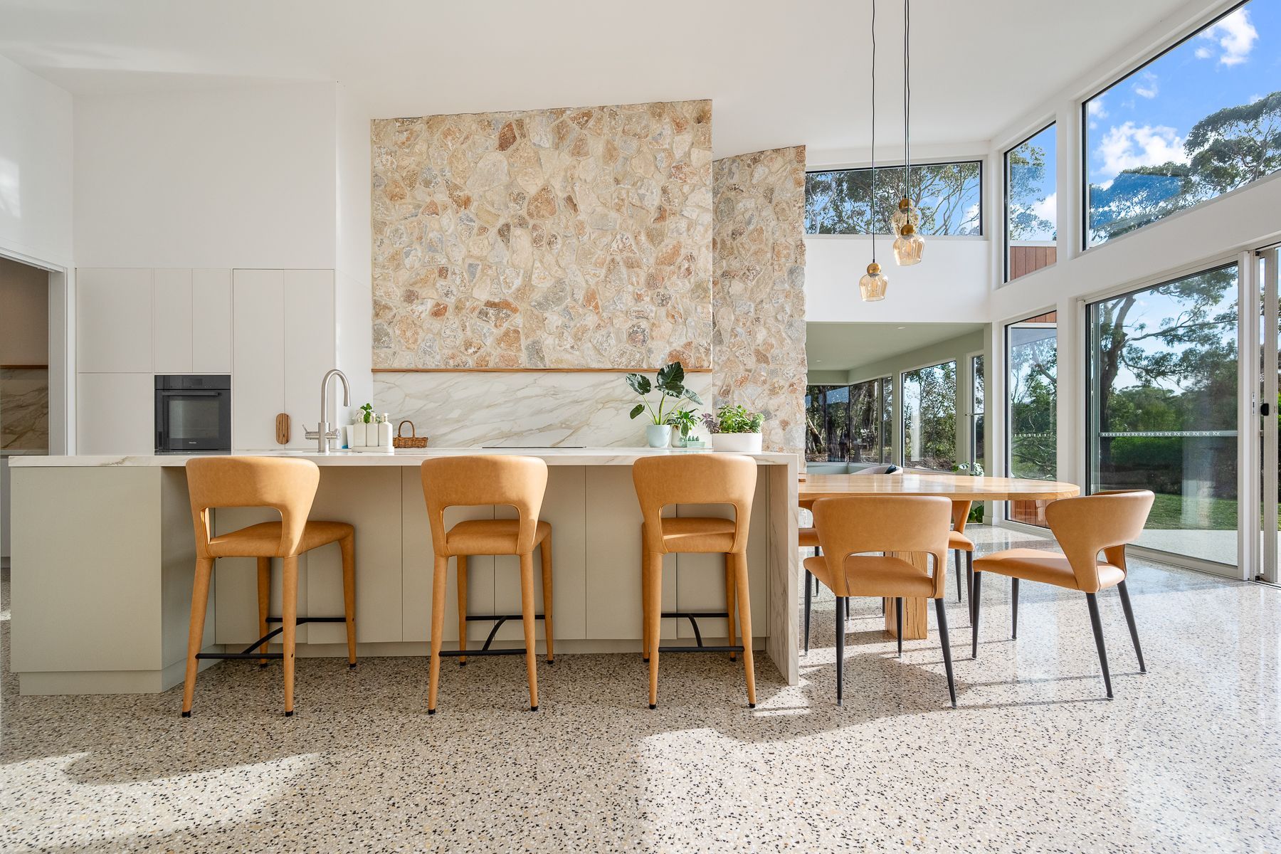 A modern kitchen with a large stone wall, light wood accents, and a dining table with outdoor views.