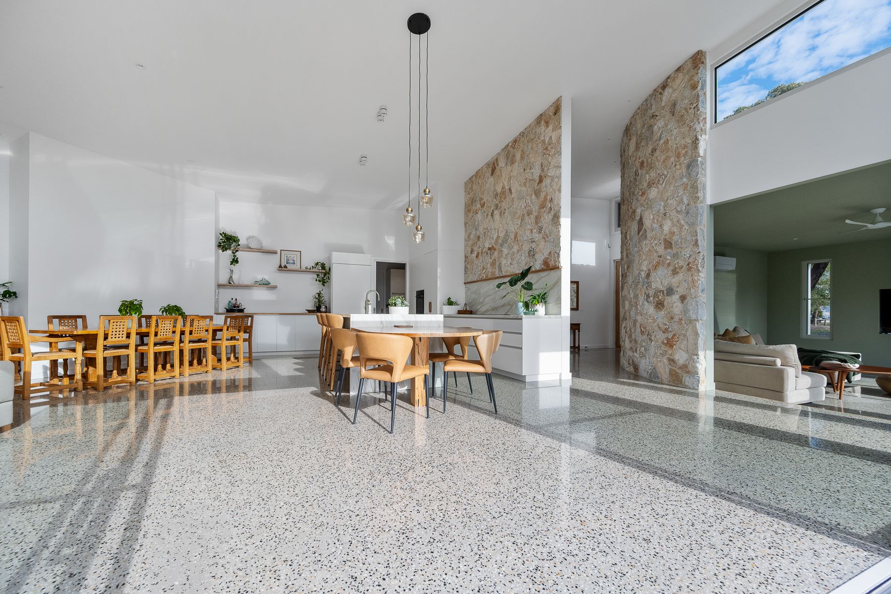 Spacious, modern interior with terrazzo floors, two dining tables, and stone accent walls. Sunlight streams in from large windows.