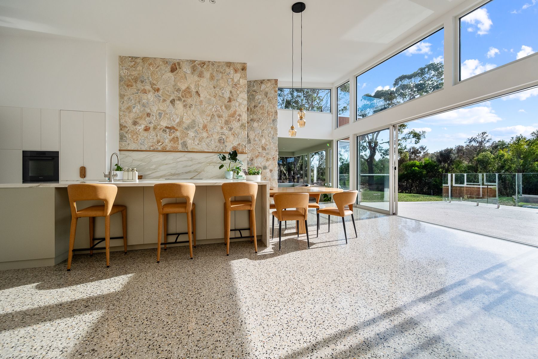 Modern kitchen with terrazzo floors and floor-to-ceiling windows. Features a stone accent wall, breakfast bar with wooden stools, and outdoor view.