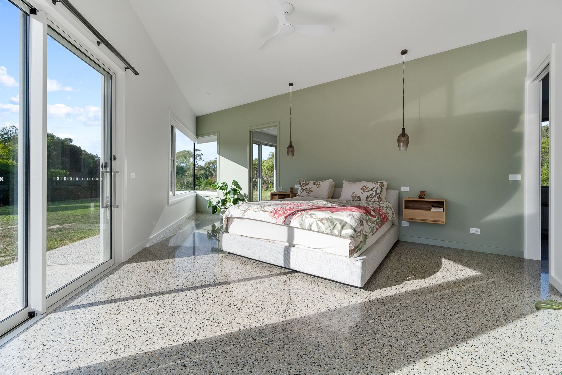 Spacious bedroom with large windows overlooking a yard, pale green accent wall behind the bed, and terrazzo flooring.