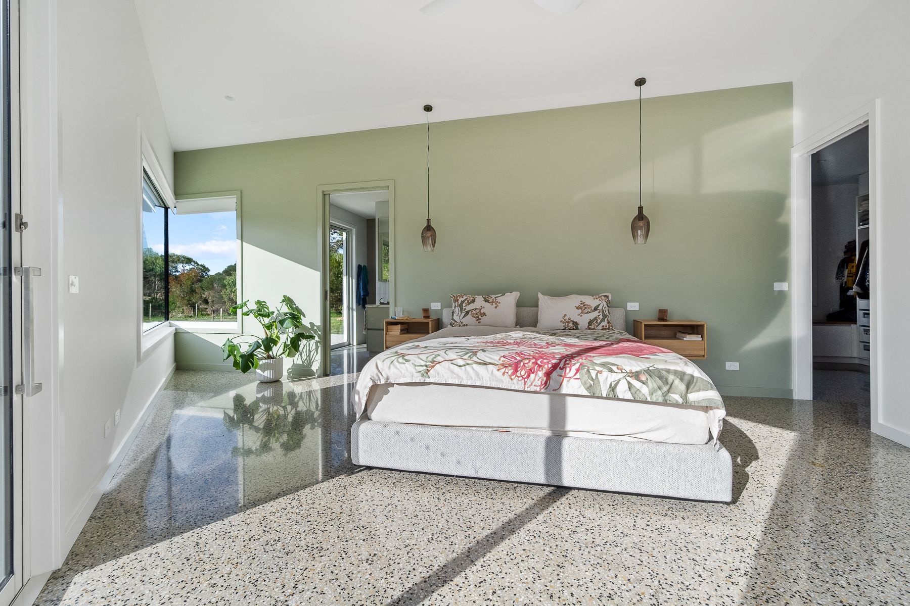 Bedroom with sage green accent wall, large window, and bed with floral bedding. Polished concrete floor reflects sunlight.