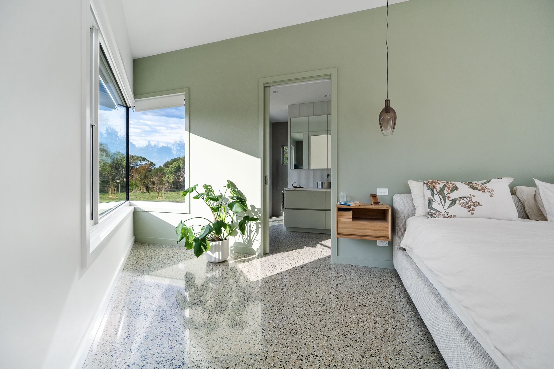 Bedroom with green walls, terrazzo floor, and a large window overlooking greenery. A bed with white bedding is on the right, a bathroom is visible in the doorway.