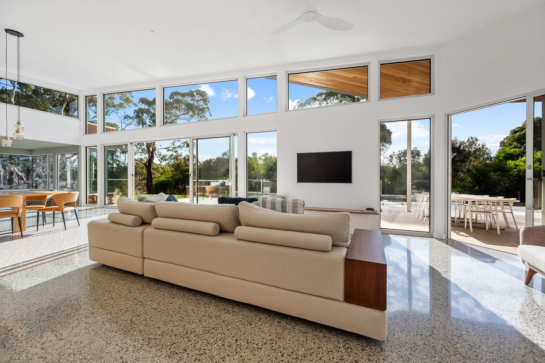 Bright, airy living room with large windows and a beige sectional sofa. Sunlight streams in, illuminating the polished stone floor and outdoor patio.