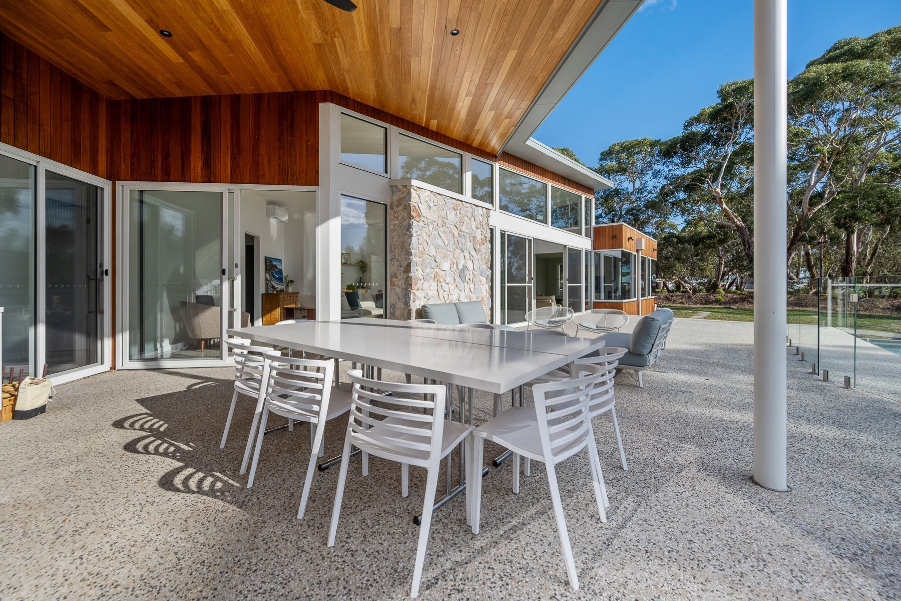 Outdoor dining area of a modern home with a large table and white chairs. The setting is a patio with a wooden ceiling and trees in the background.