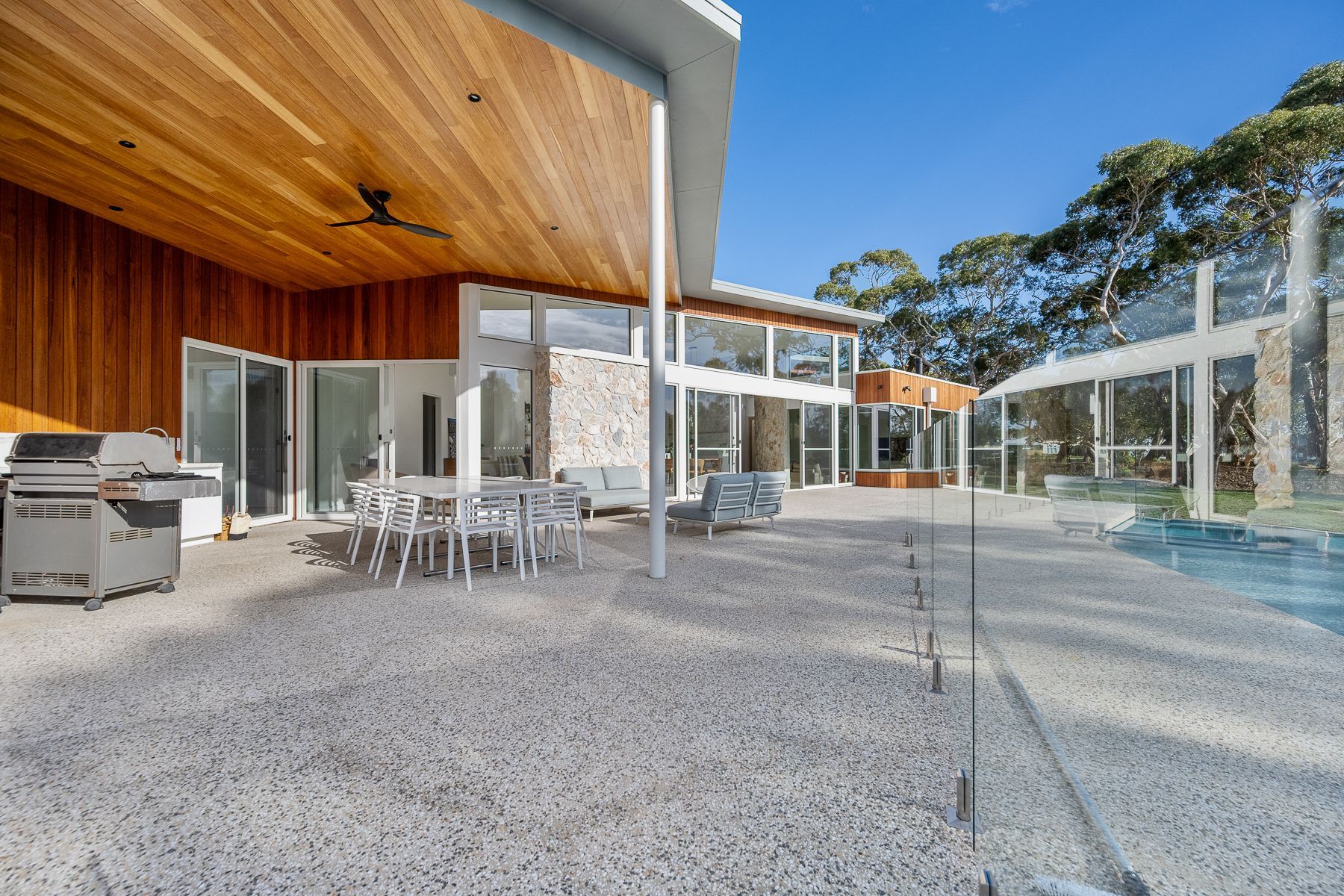Patio with outdoor dining set and grill, attached to a modern house with floor-to-ceiling windows, overlooking a wooded area.