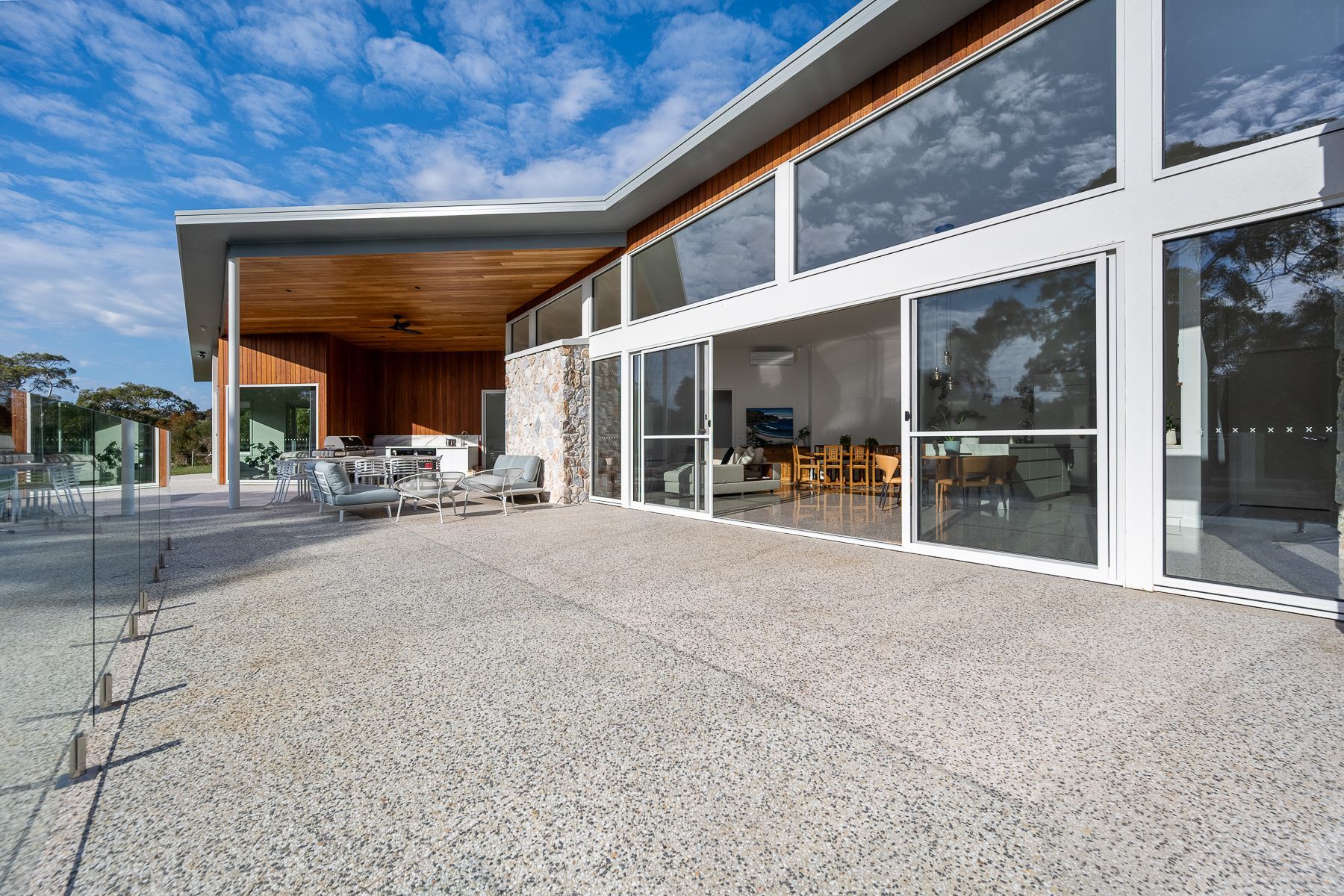 Spacious modern building with large windows, patio, and light-colored concrete flooring. Glass railings define the outdoor area under a clear blue sky.