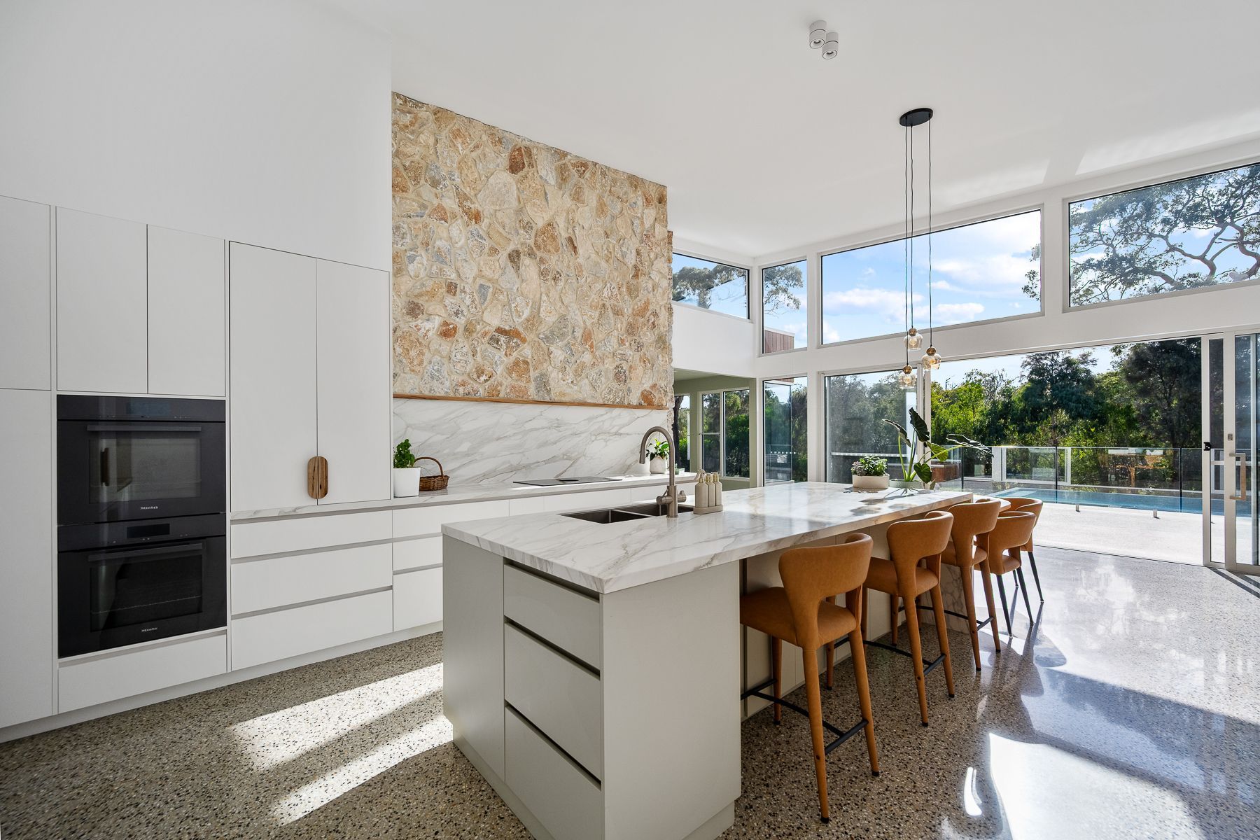 Modern kitchen with white cabinets, a marble countertop, a large stone backsplash, and a central island with brown bar stools. Large windows overlook a pool.