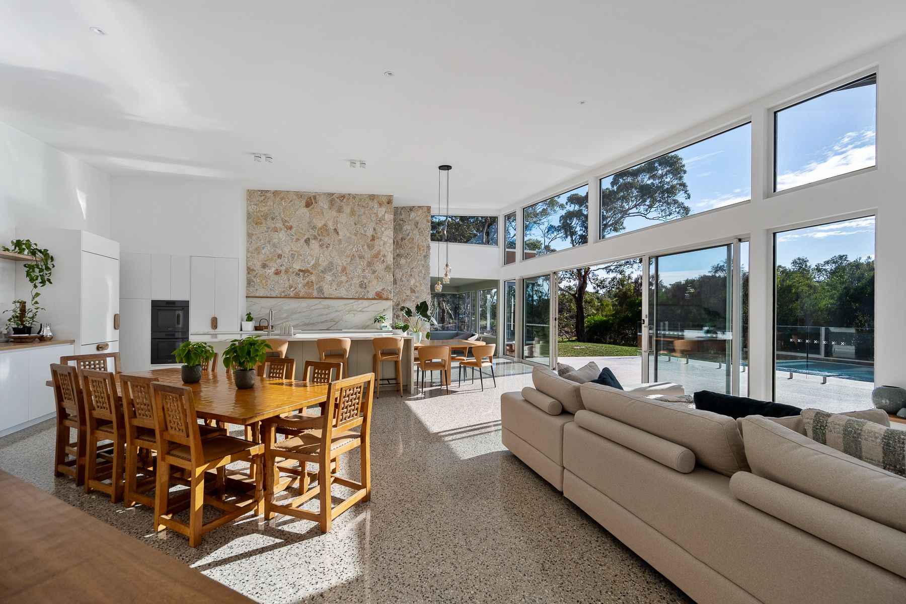 Spacious, bright living area with large windows overlooking a pool and trees. It features a dining area, kitchen, and a large sofa.