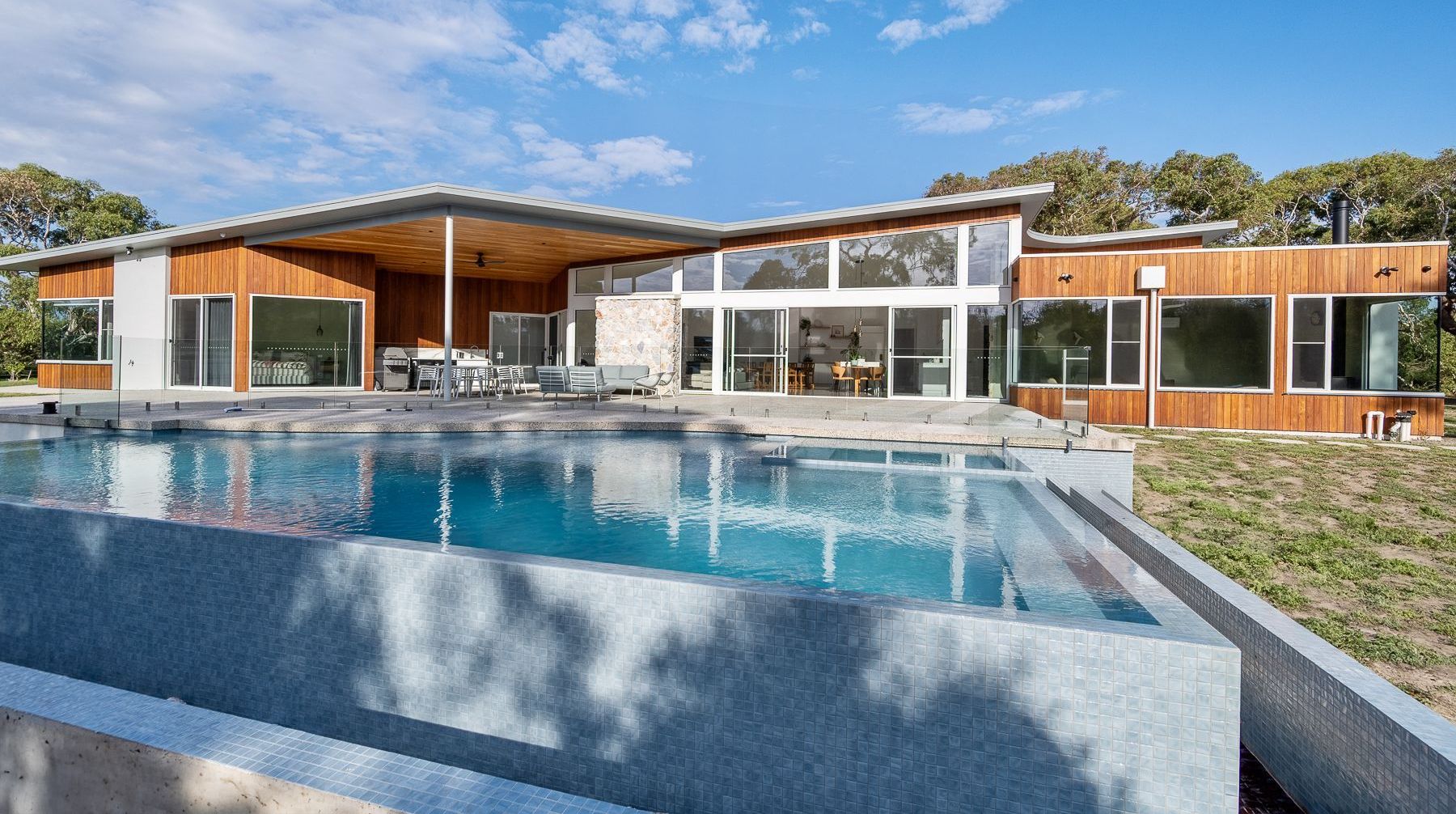 Modern home with a long, rectangular pool, wooden siding, and floor-to-ceiling windows on a sunny day.