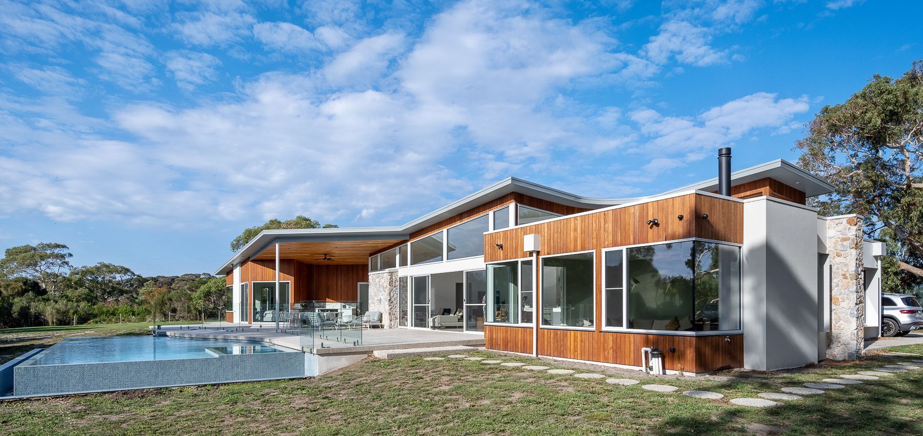 Modern house with wood and stone siding, large windows, and a pool, set against a blue sky.