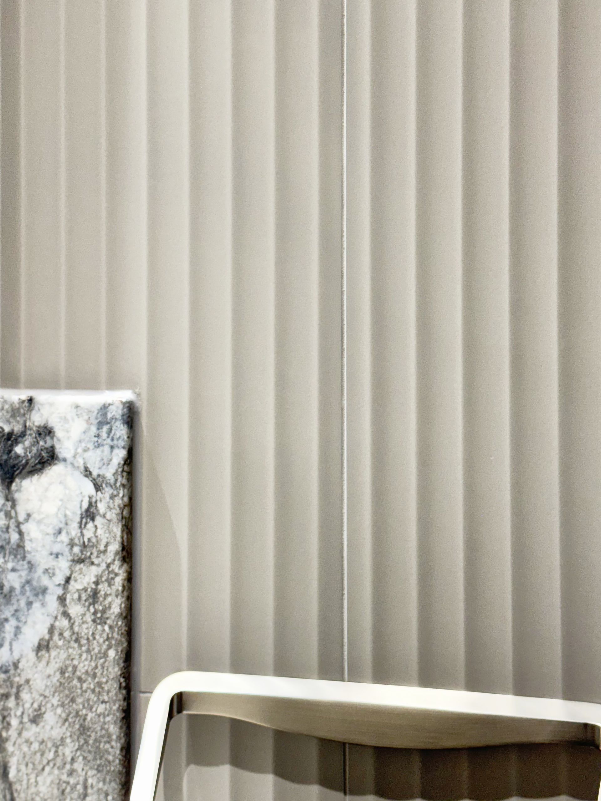 Vertical blinds, light gray, against a wall; an abstract painting and part of a white chair on the left.