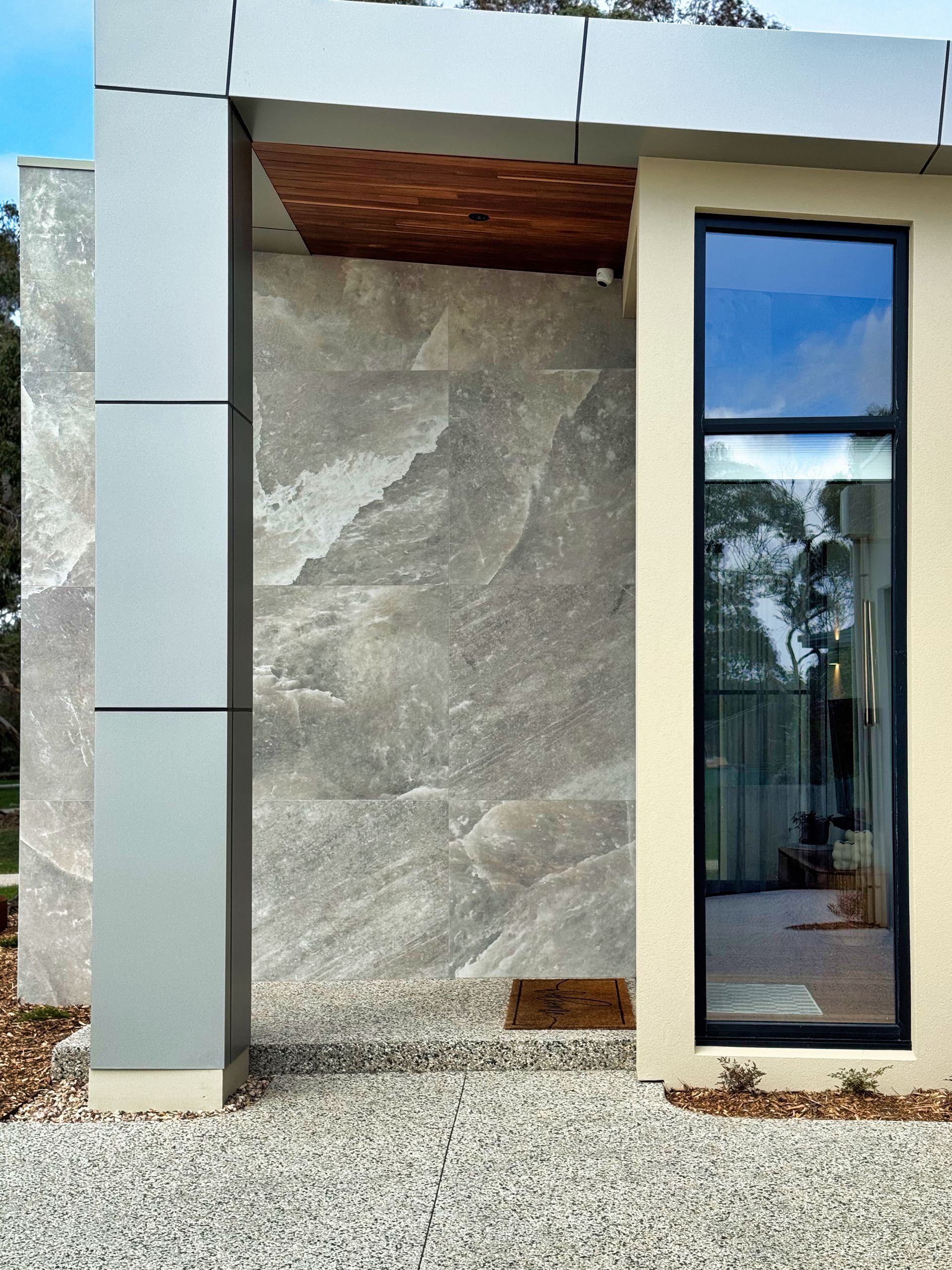 Modern building exterior with large grey stone wall, narrow black-framed window, and gravel ground.