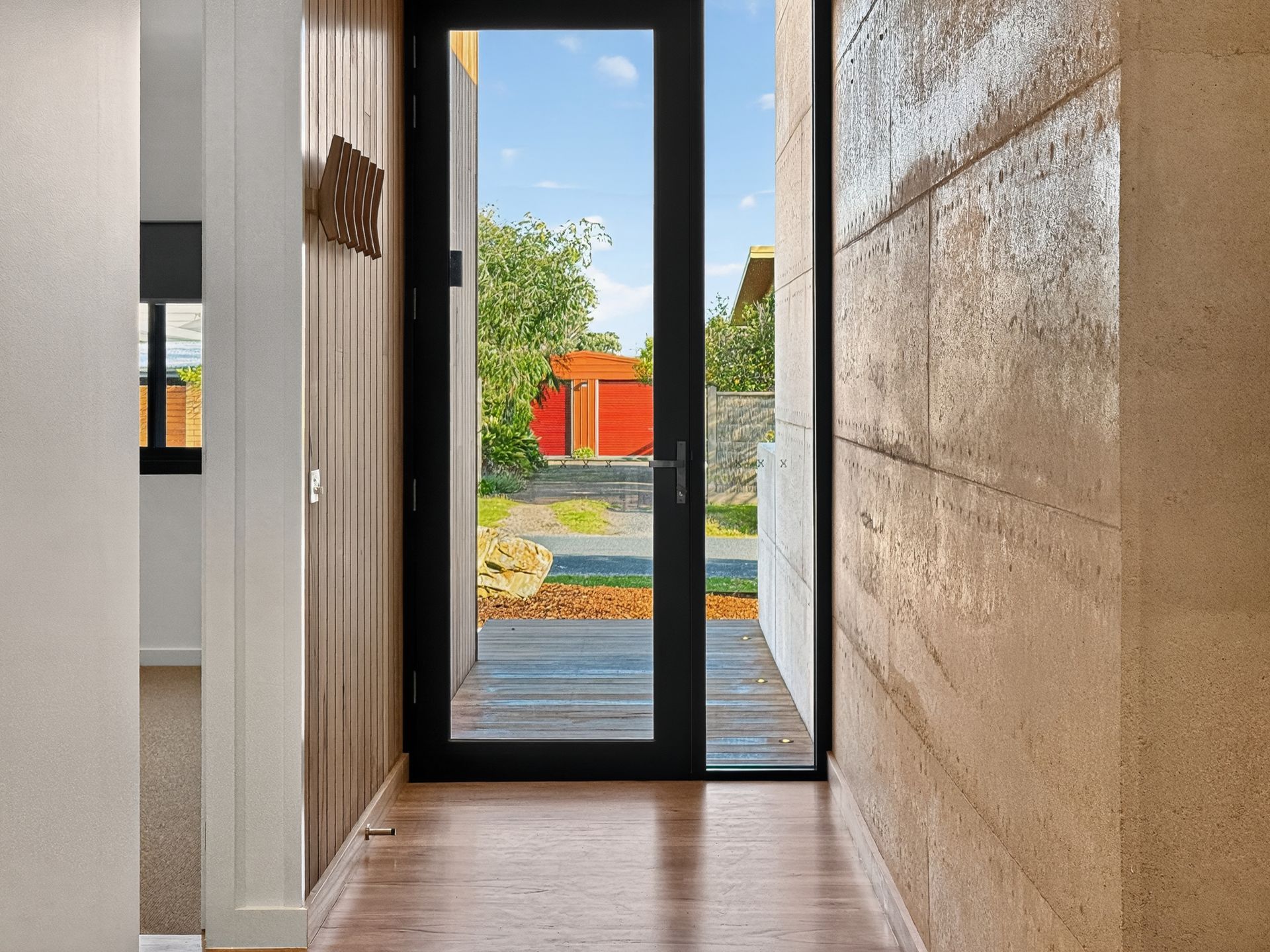 Hallway with view of yard through black-framed door. Brown walls, wood floor, and blue sky.