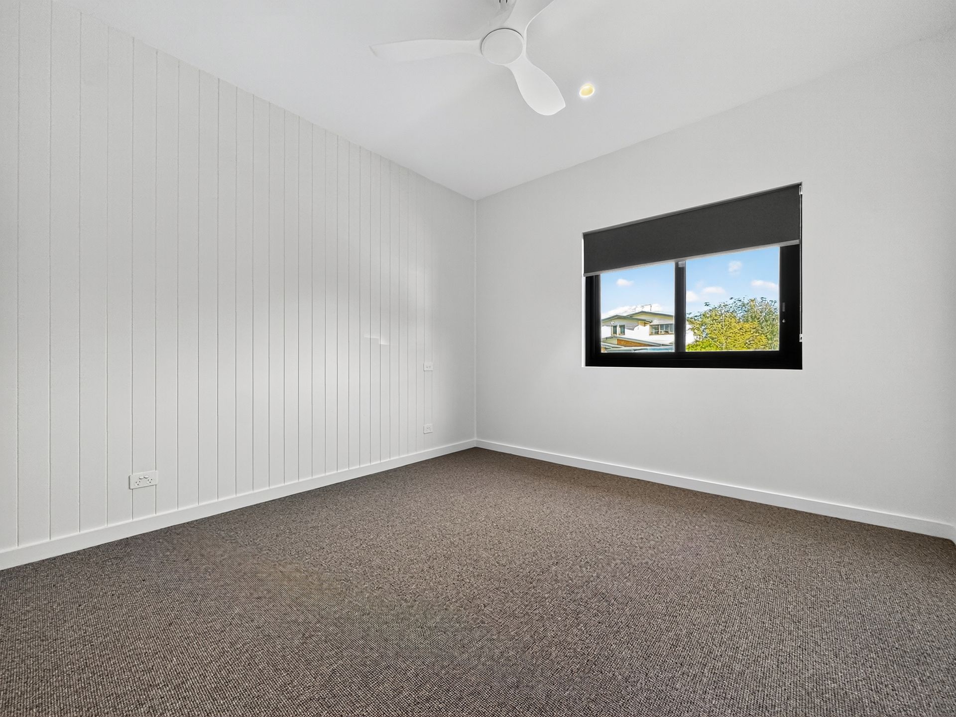 Empty bedroom with white paneled wall, dark carpet, window with shade, and ceiling fan.