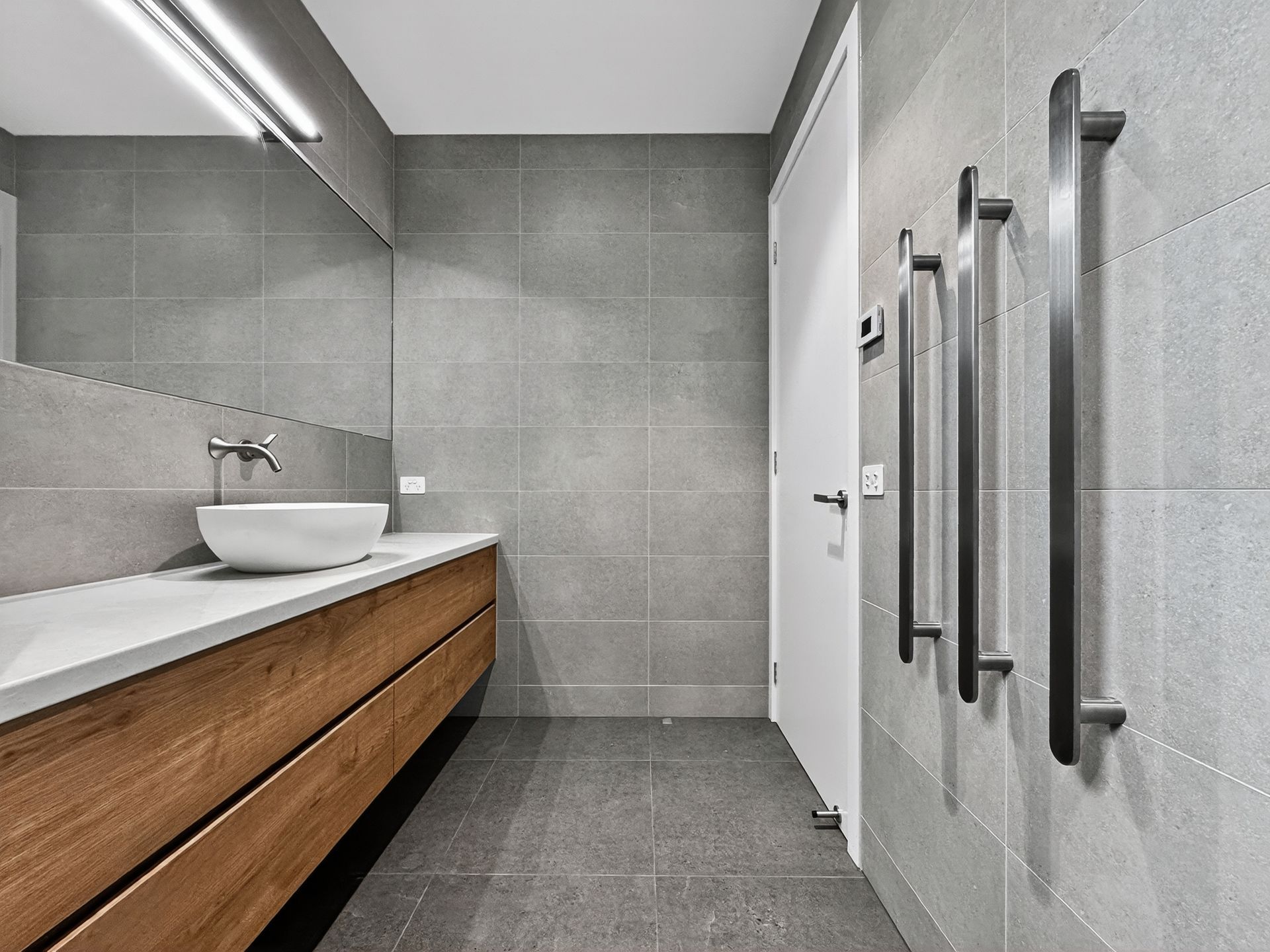 Modern bathroom with gray tile walls and floor, wood vanity, white sink, and towel warmers.