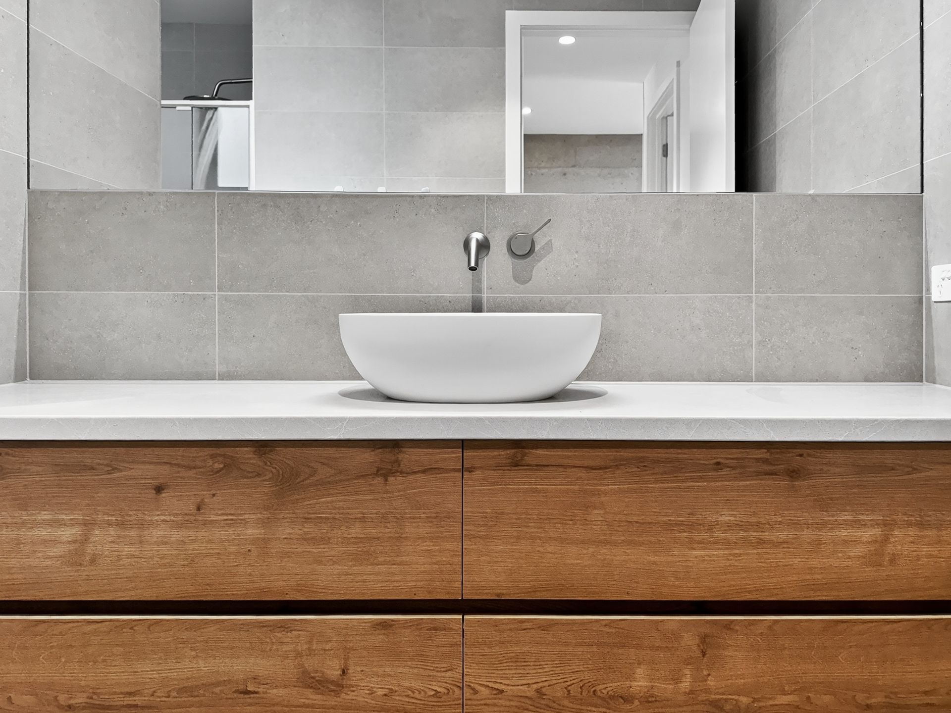Bathroom vanity with a wood-grain cabinet, white countertop, and vessel sink. Gray tiled wall and large mirror.