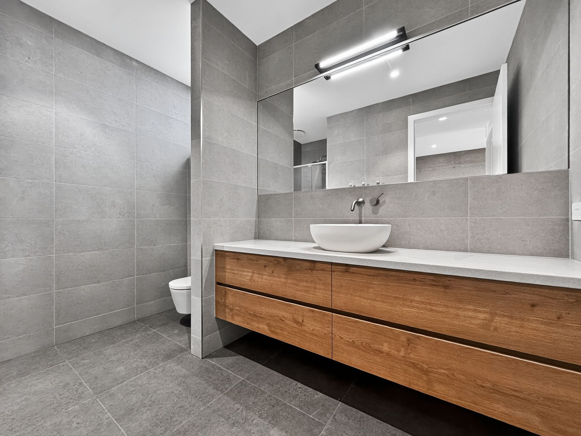 Modern bathroom with gray tiled walls and floor, wood vanity, vessel sink, and large mirror.