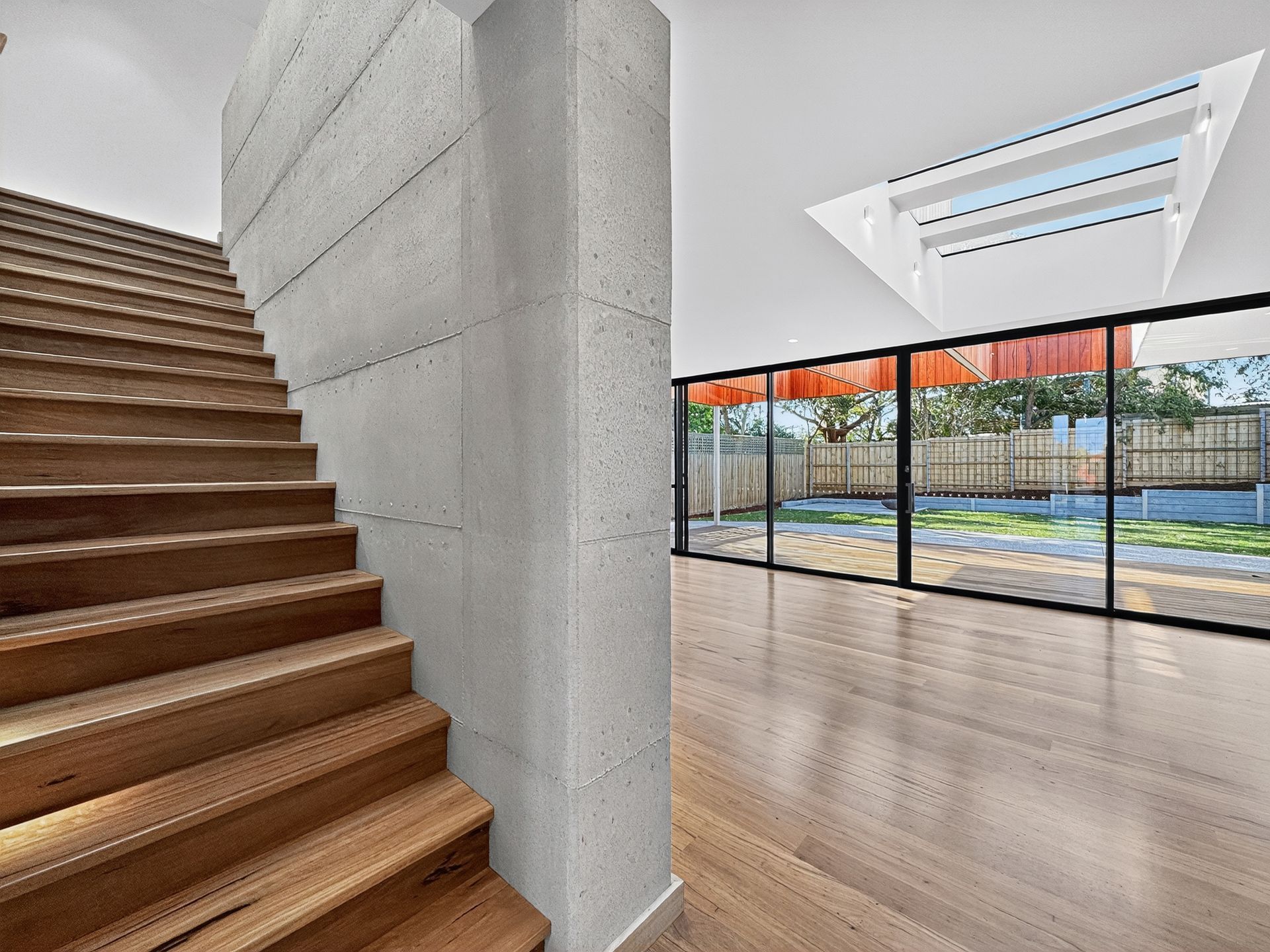 Wooden staircase next to concrete pillar; bright room with large windows and backyard view.
