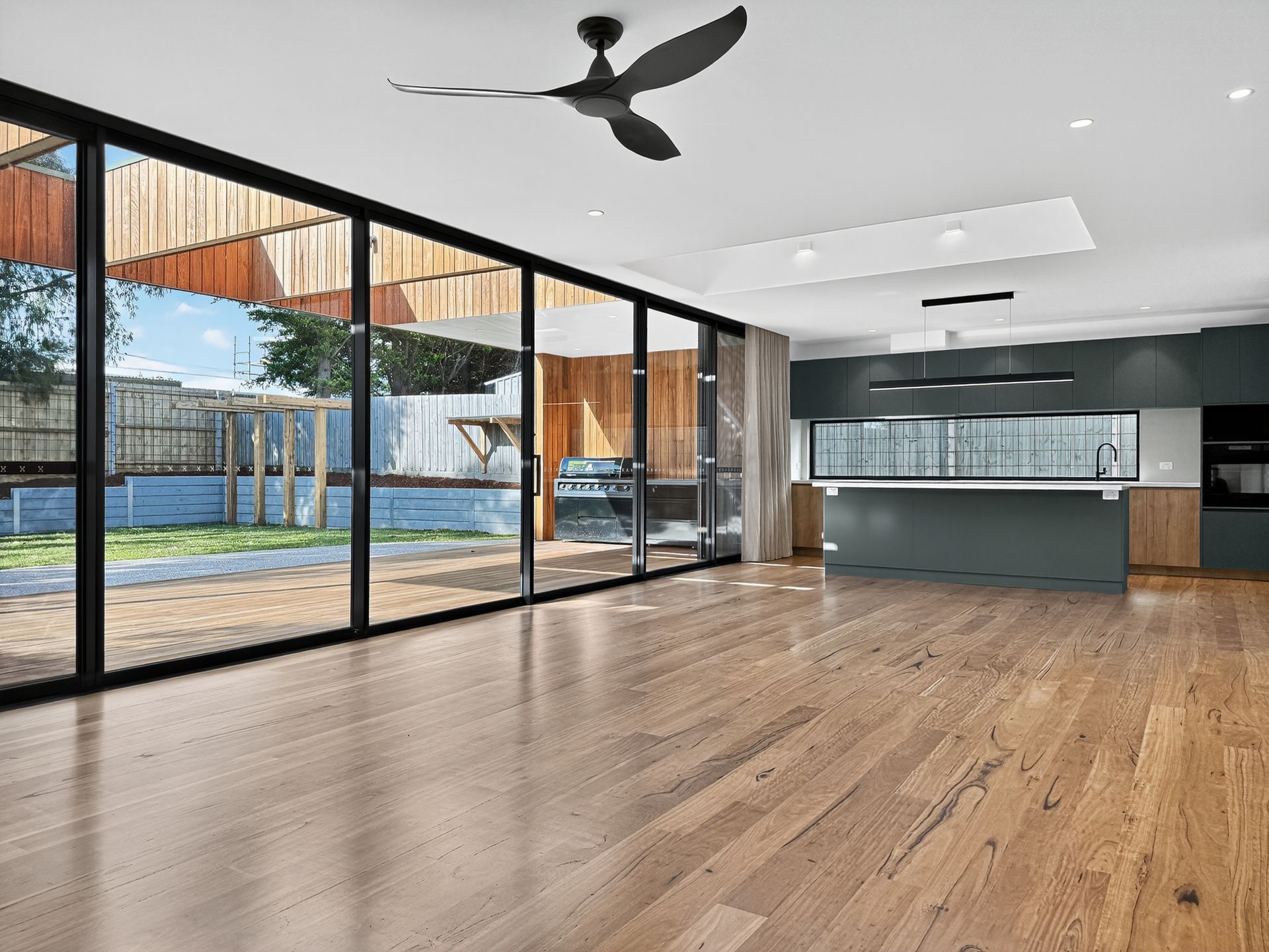 Modern living room with large glass doors, wooden floors, and a dark teal kitchen.