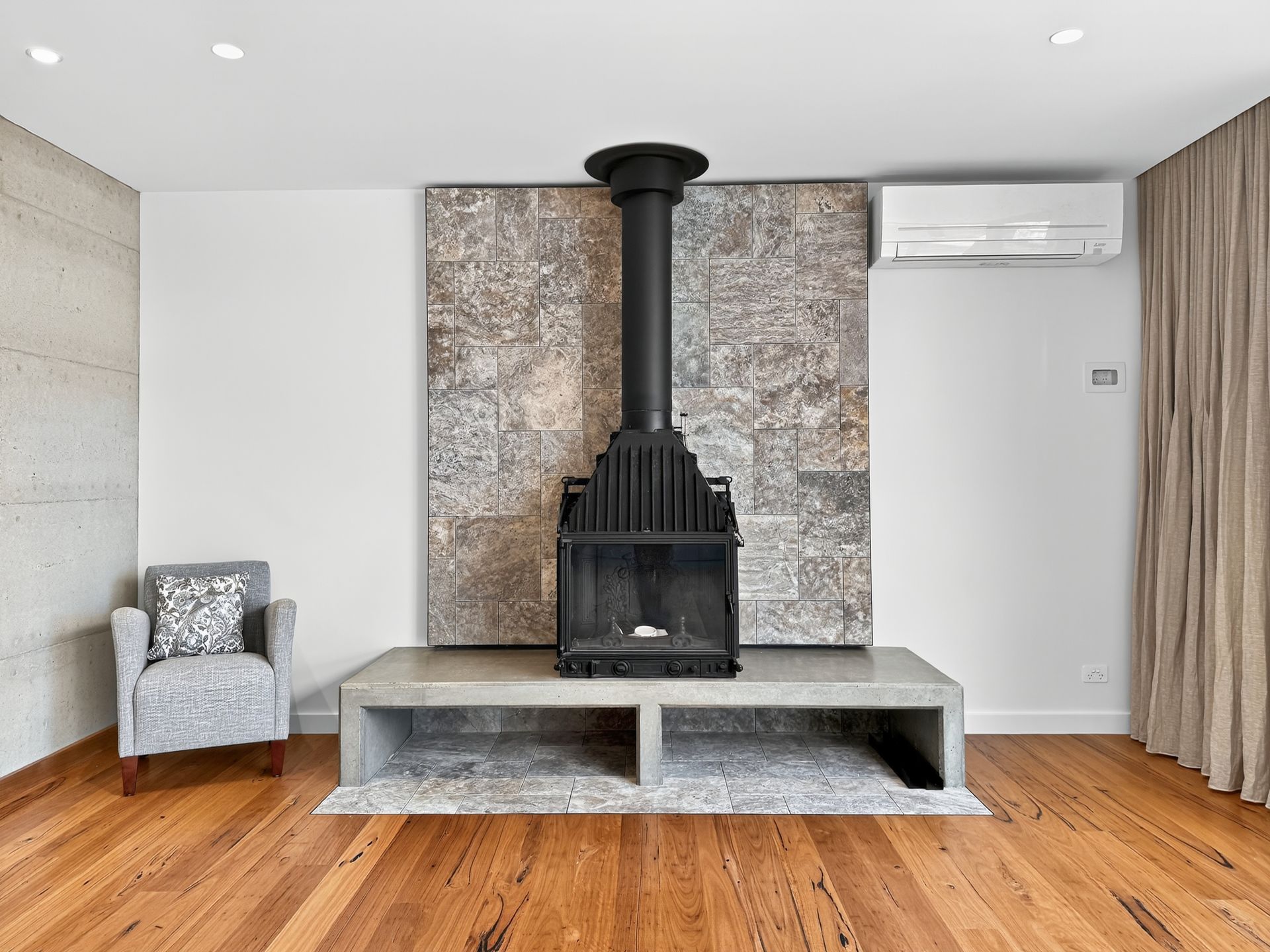 Fireplace with black stove, concrete hearth, tiled backdrop, wood floor, and armchair.