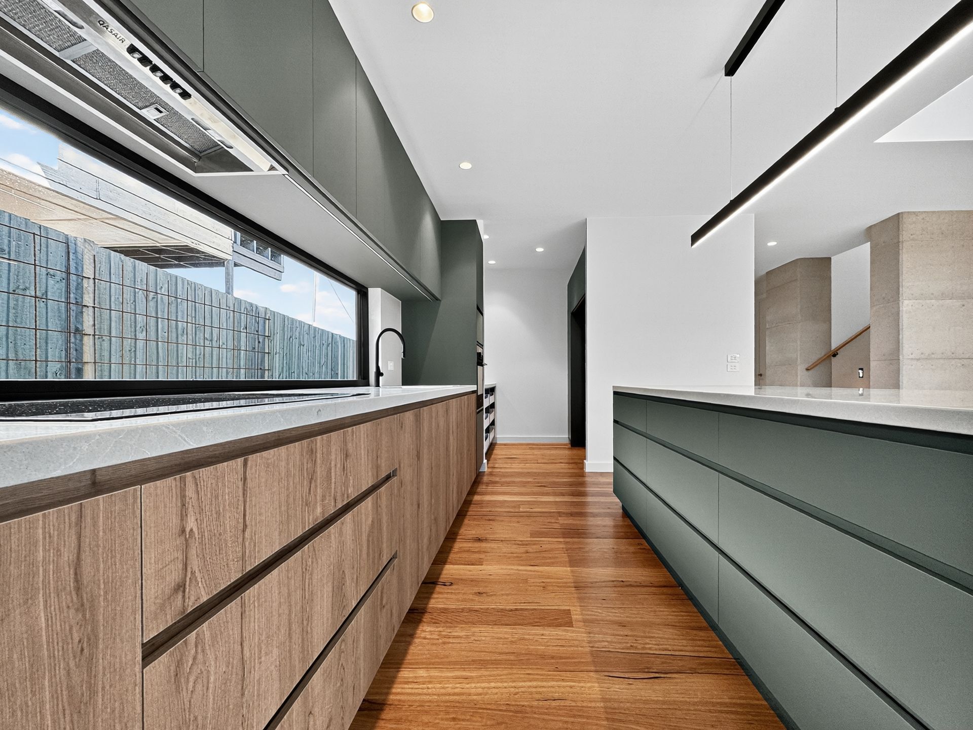 Modern kitchen with wooden and green cabinetry, long countertop, large window, and hardwood floor.