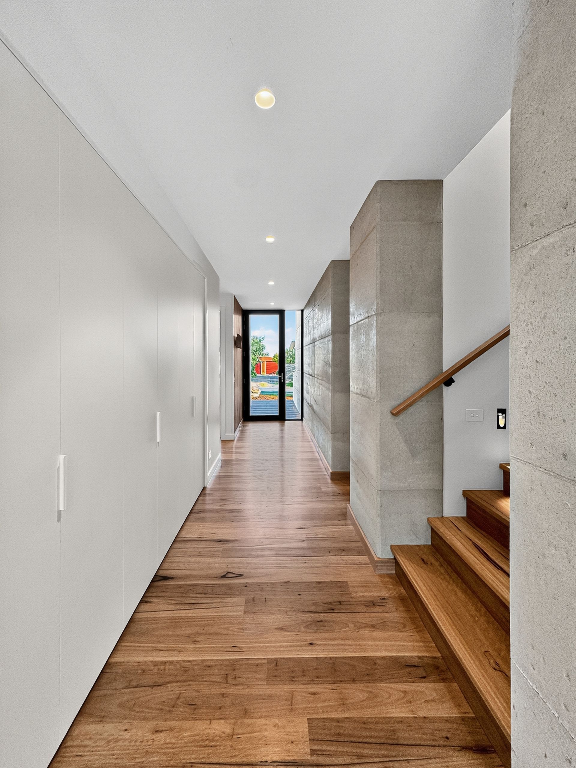 Hallway with wooden floor, white wall with cabinets, concrete pillars, and stairs leading up.