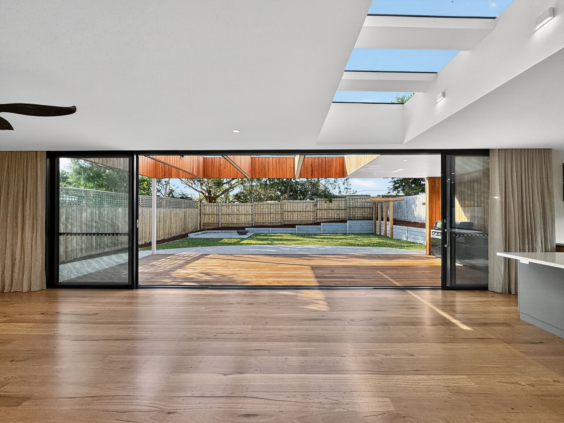 Spacious interior with open glass doors leading to a backyard. Wooden floors, skylights, and curtains are visible.