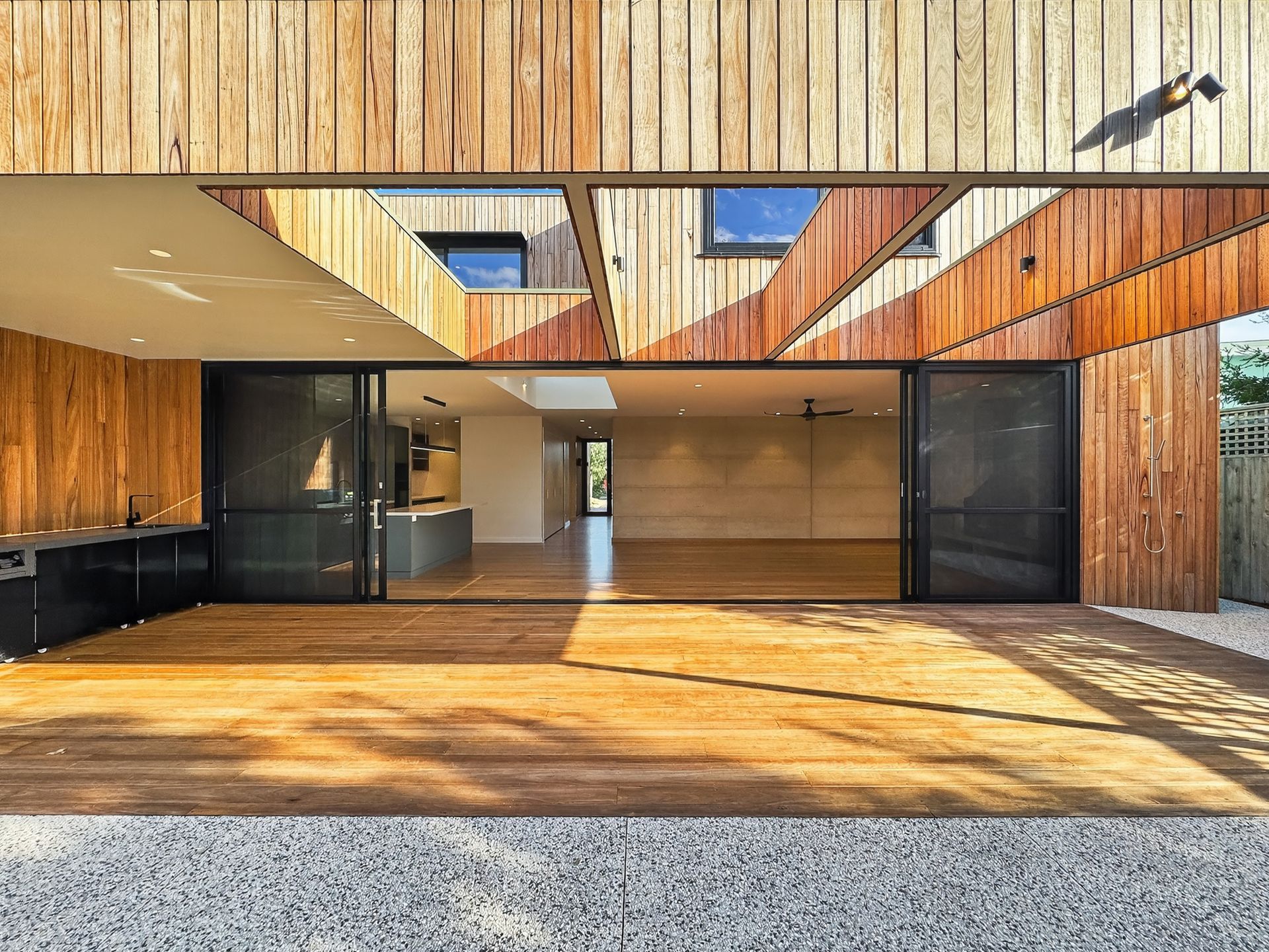 Wooden house exterior with open sliding doors, revealing kitchen and interior. Sunny, spacious.