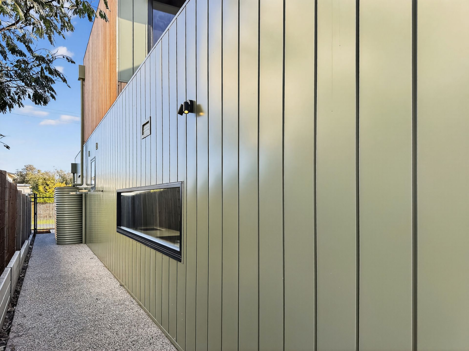 Side of a modern building with green metal siding and a narrow gravel path.