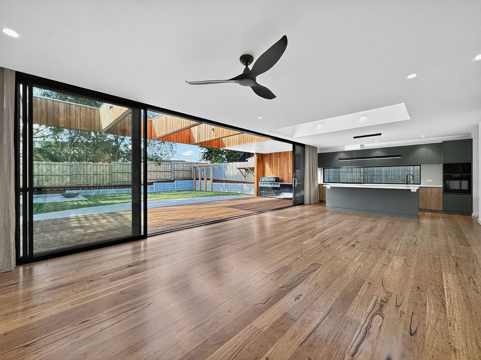 Spacious interior with open sliding doors to a backyard; wooden floor, modern kitchen, and ceiling fan.