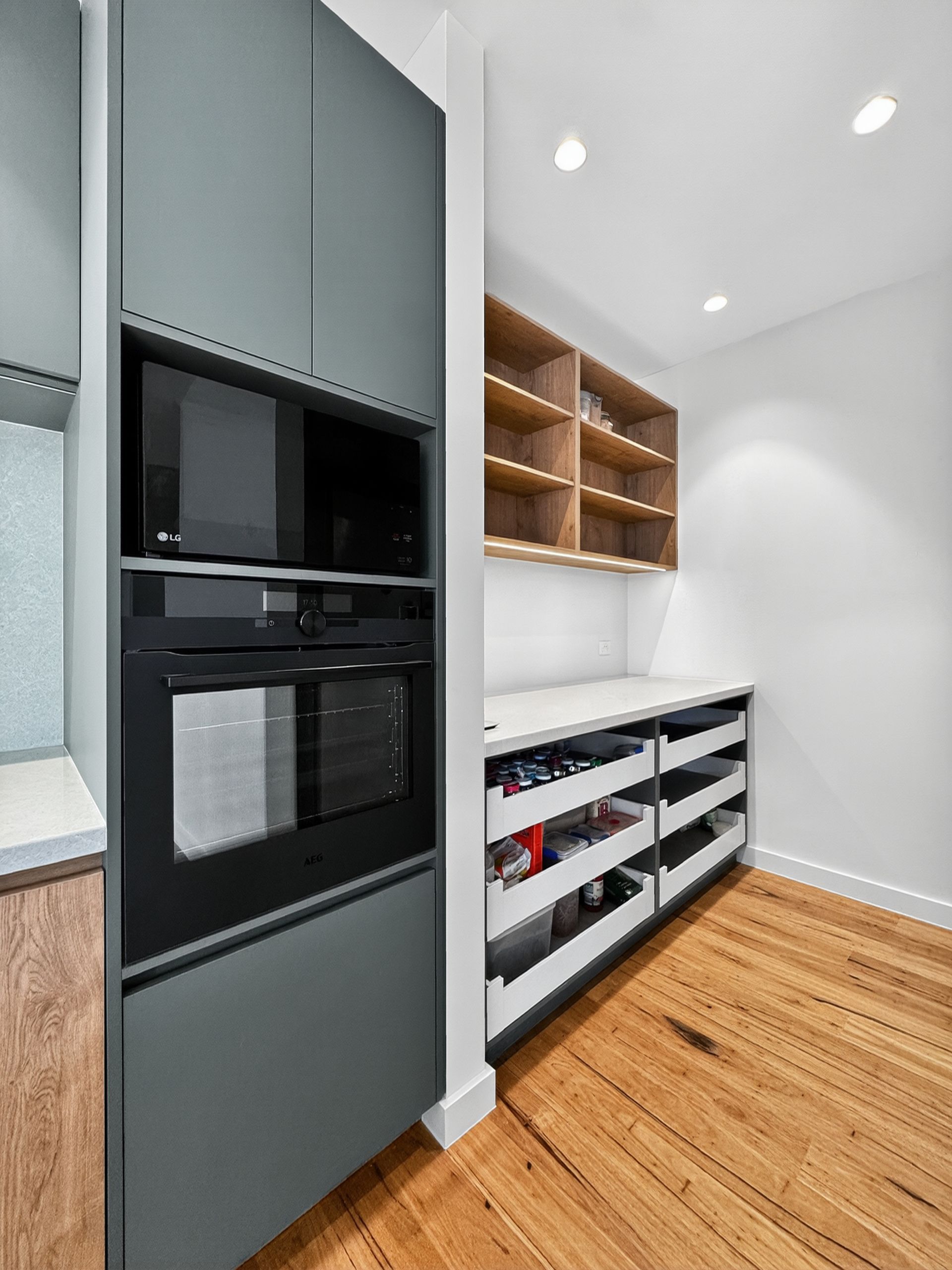 Built-in oven and microwave in a gray cabinet, with wooden pantry shelves and drawers, and hardwood floor.