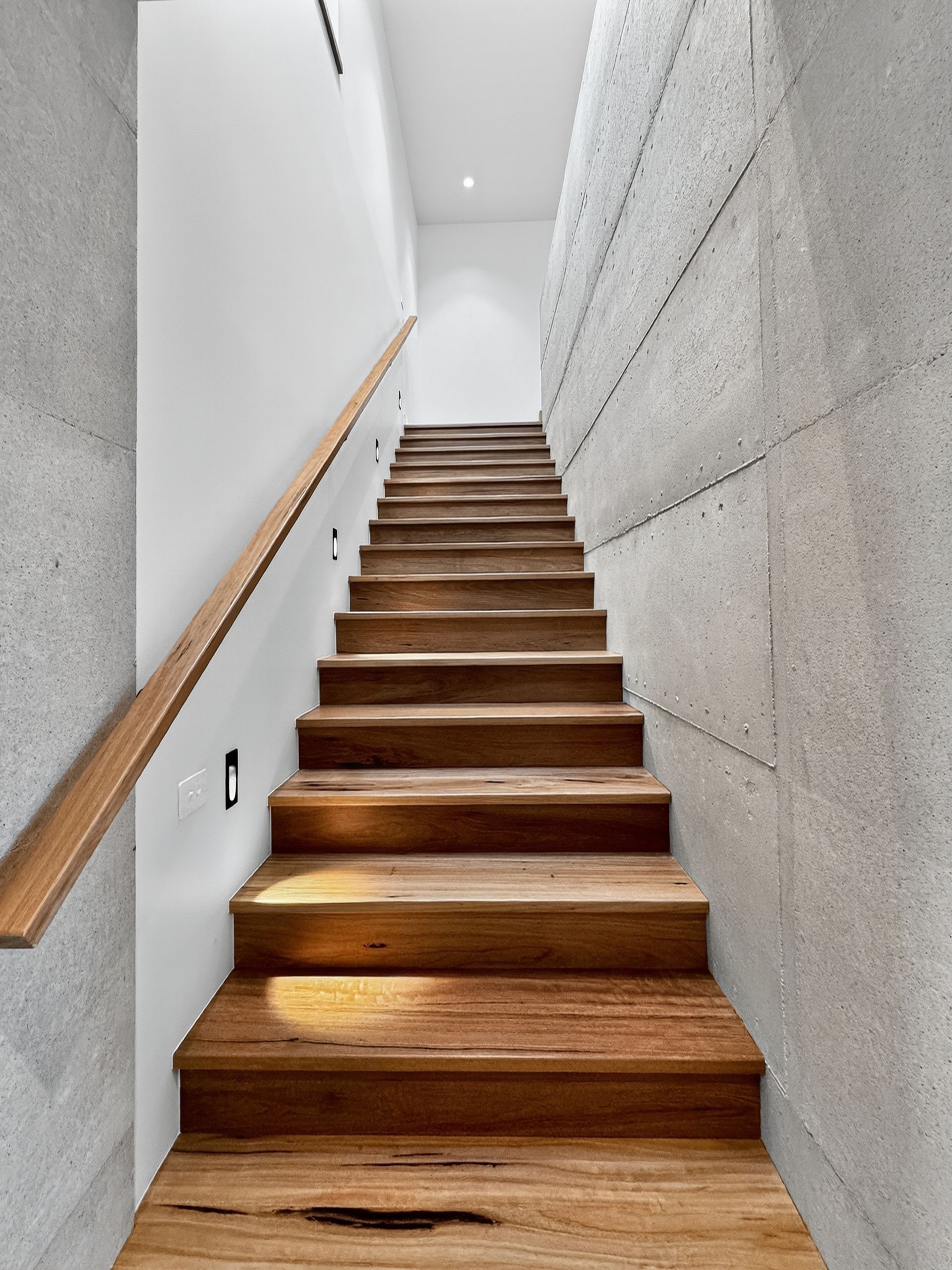 Wooden staircase with wooden handrail ascending between white and textured gray walls.