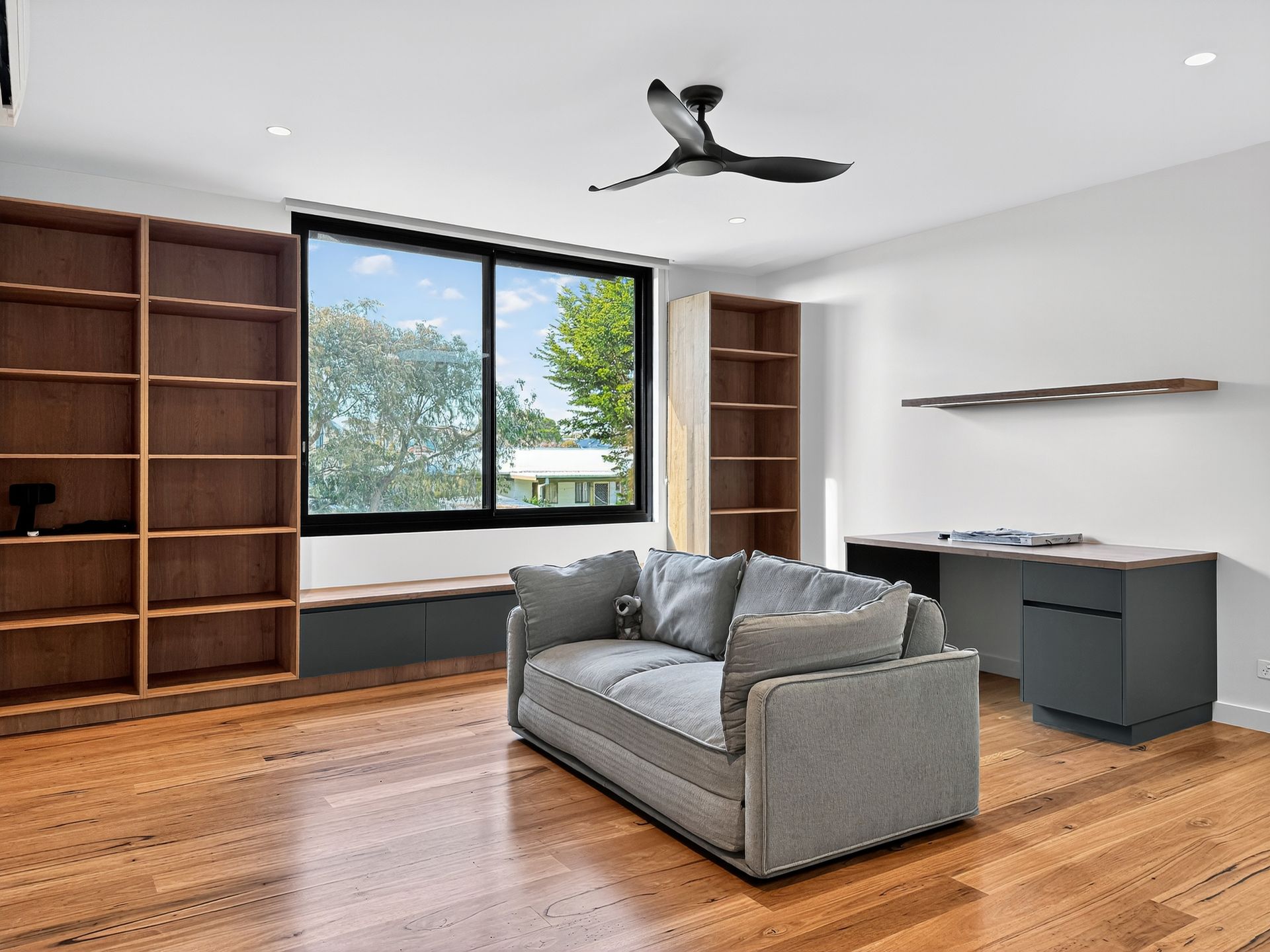 Spacious room with wood floors, built-in bookshelves, a couch, desk, and window overlooking trees.
