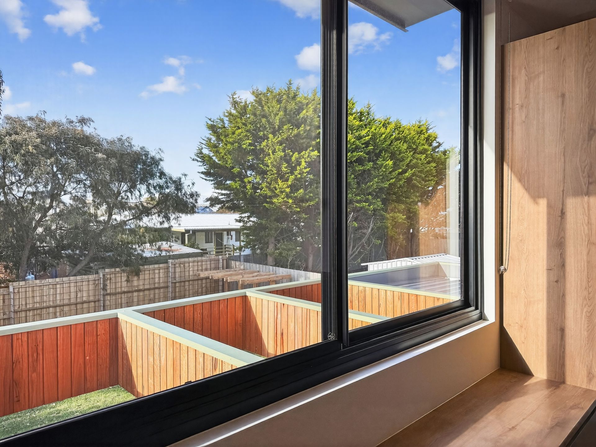 Interior view through a large window overlooking a wooden fence, trees, and sky. The window frame is black.
