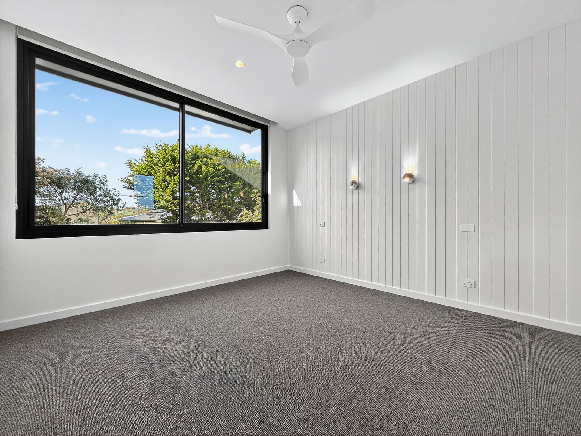Empty room with a large window and white paneling. Carpet flooring, ceiling fan, two wall sconces.