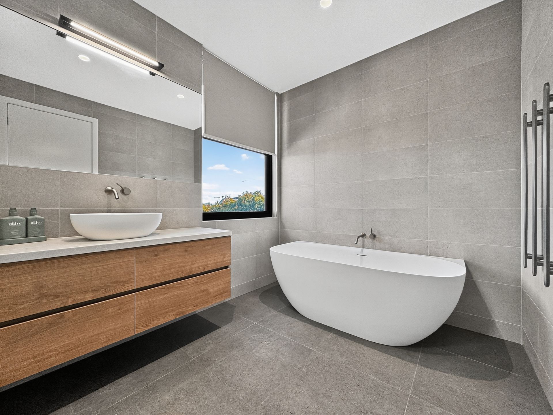 Modern bathroom with a white tub, wooden vanity, gray tile, and a window.